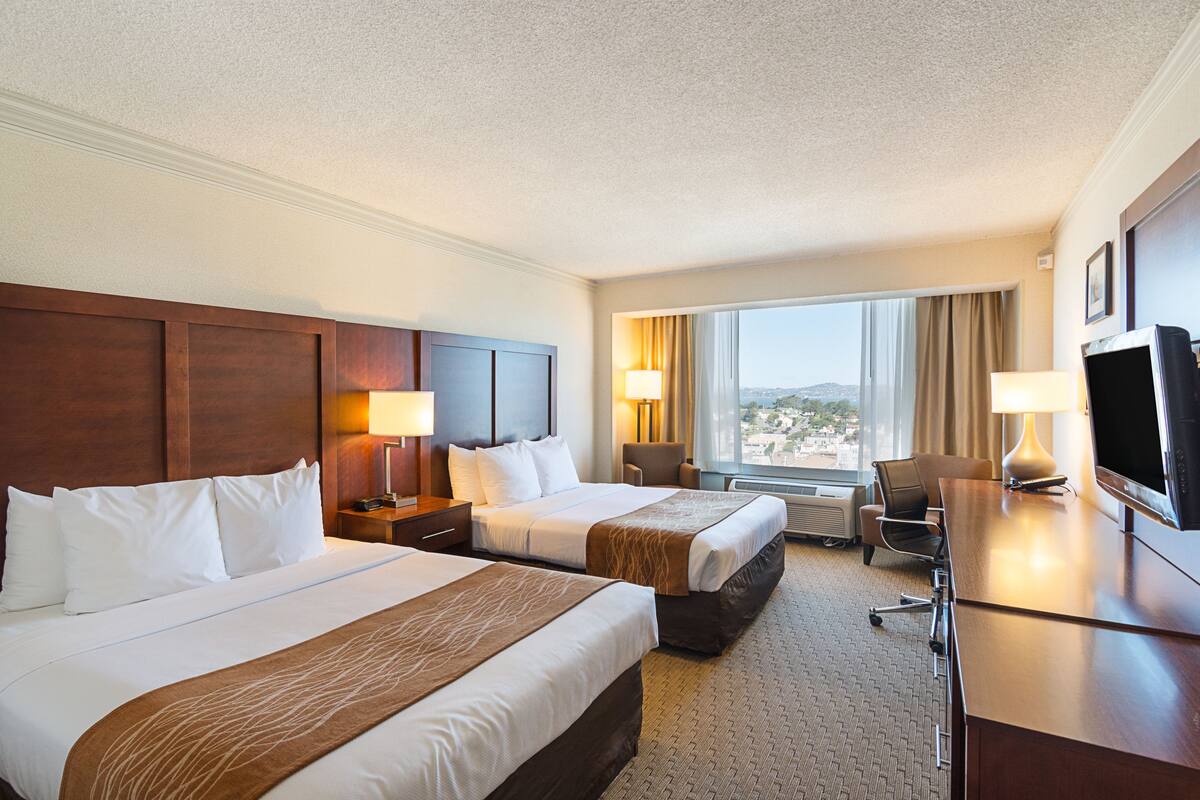 A hotel room with two beds, a desk, a TV, and a view of the city.