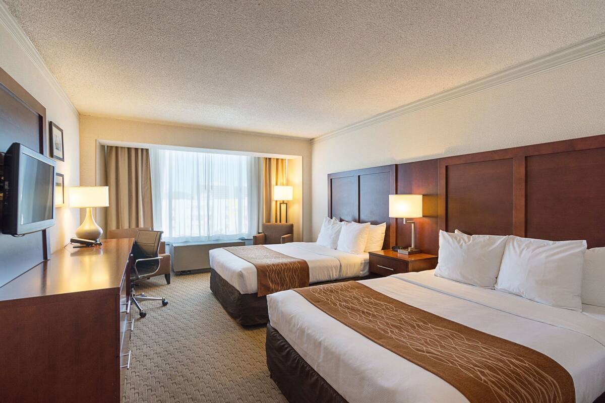 A hotel room with two beds, a desk with a TV, a chair, a desk lamp, and a window with curtains.