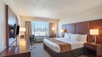 A hotel room with a large bed, two bedside tables with lamps, a desk with a chair, a TV, and a view of the city through large windows.