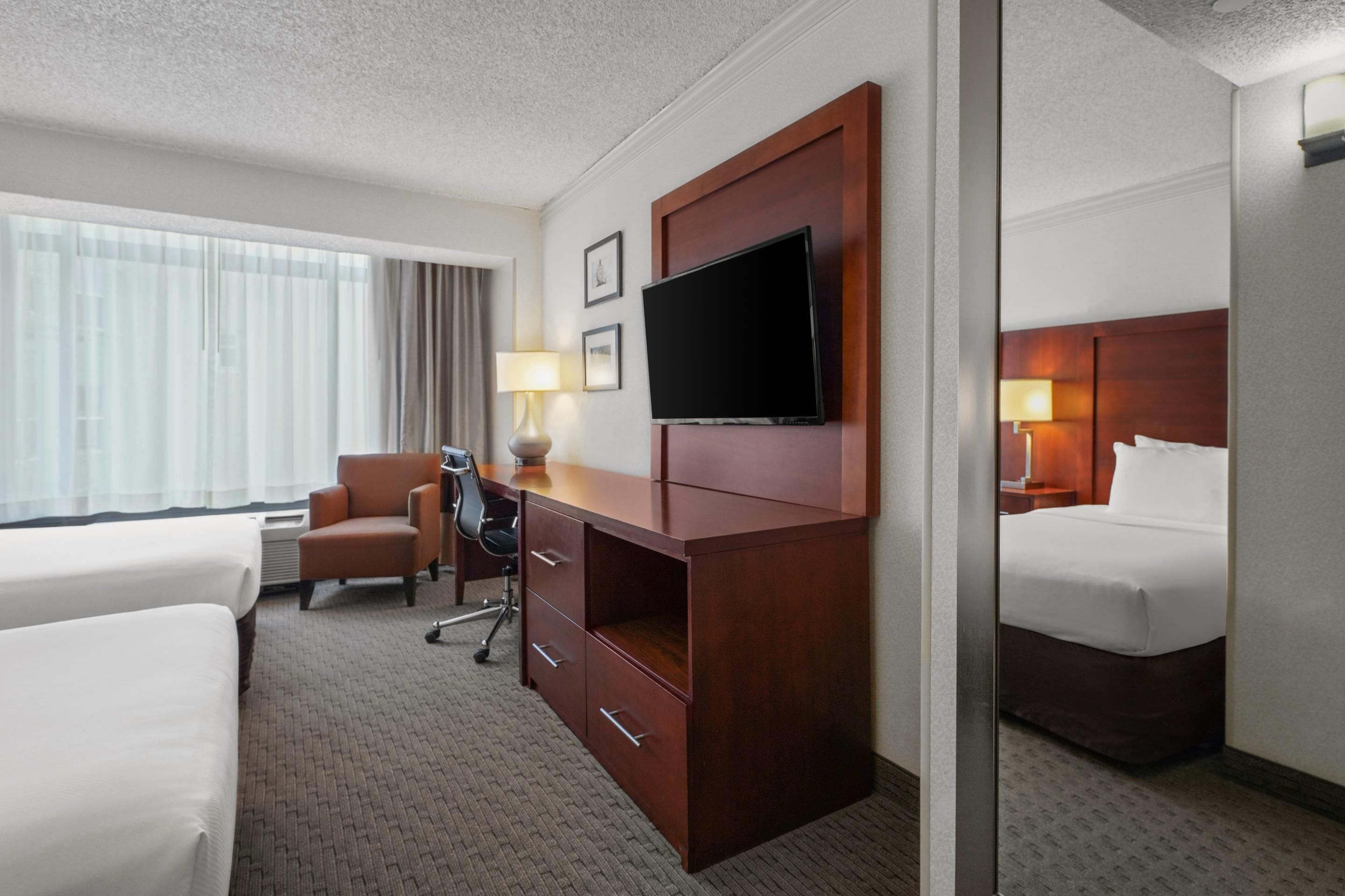 A hotel room with two beds, a desk with a chair, a television, and a large window with curtains.