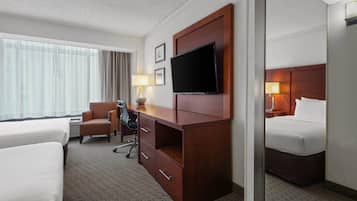 A hotel room with two beds, a desk with a chair, a television, and a large window with curtains.