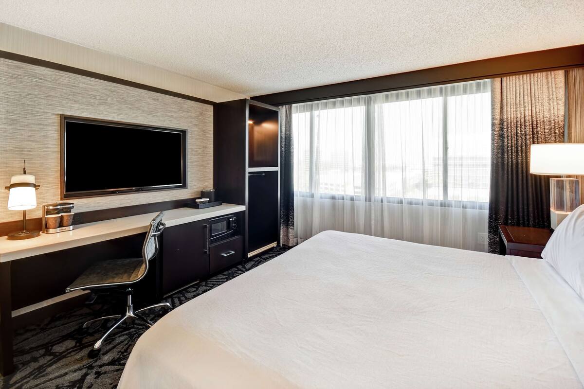 A hotel room with a large bed, a desk with a flat-screen TV, a chair, a minibar, and a window with curtains.
