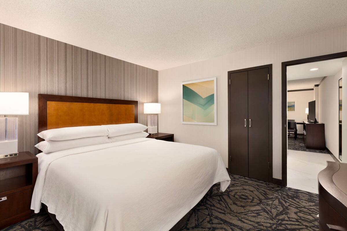 A hotel room with a large bed, a wooden headboard, a nightstand with a lamp, a wardrobe, and a carpeted floor.