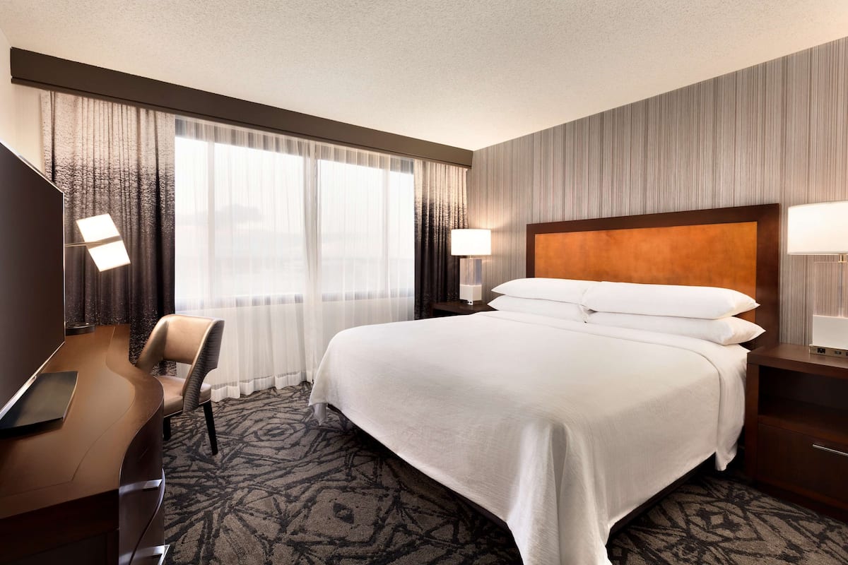 A hotel room with a large bed, a desk with a chair, a TV, and a window with curtains.