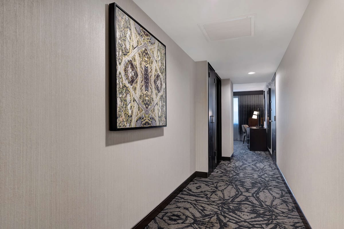 A hotel hallway with a patterned carpet, a large abstract artwork on the wall, and a door leading to a room with a desk and chair.