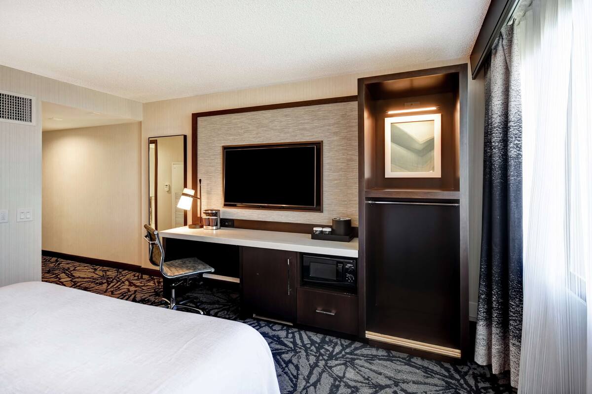 A hotel room with a bed, a desk with a flat-screen TV, a microwave, and a minibar.