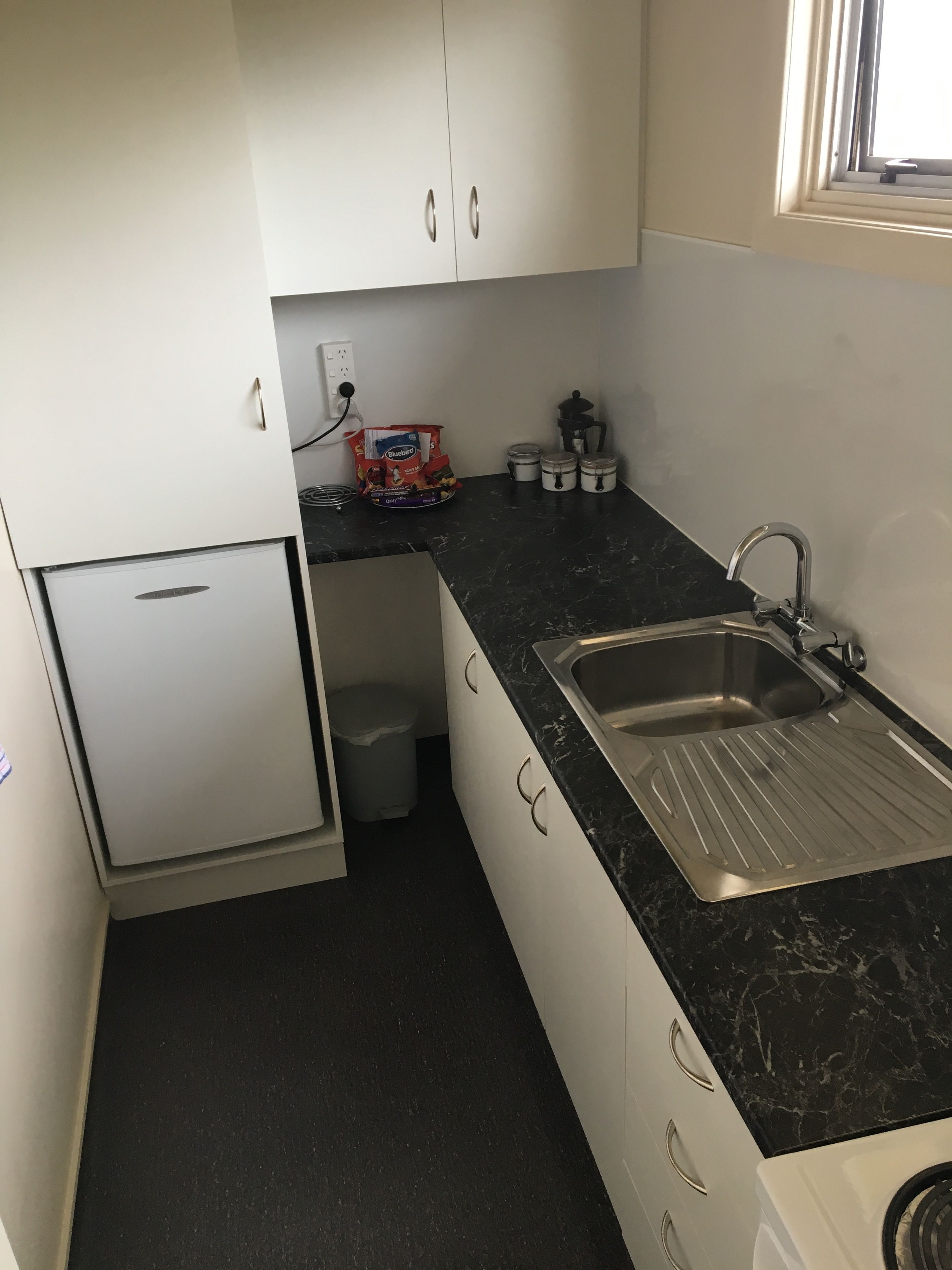 Uma cozinha compacta com armários brancos, bancada preta, pia de aço inoxidável e uma geladeira pequena.