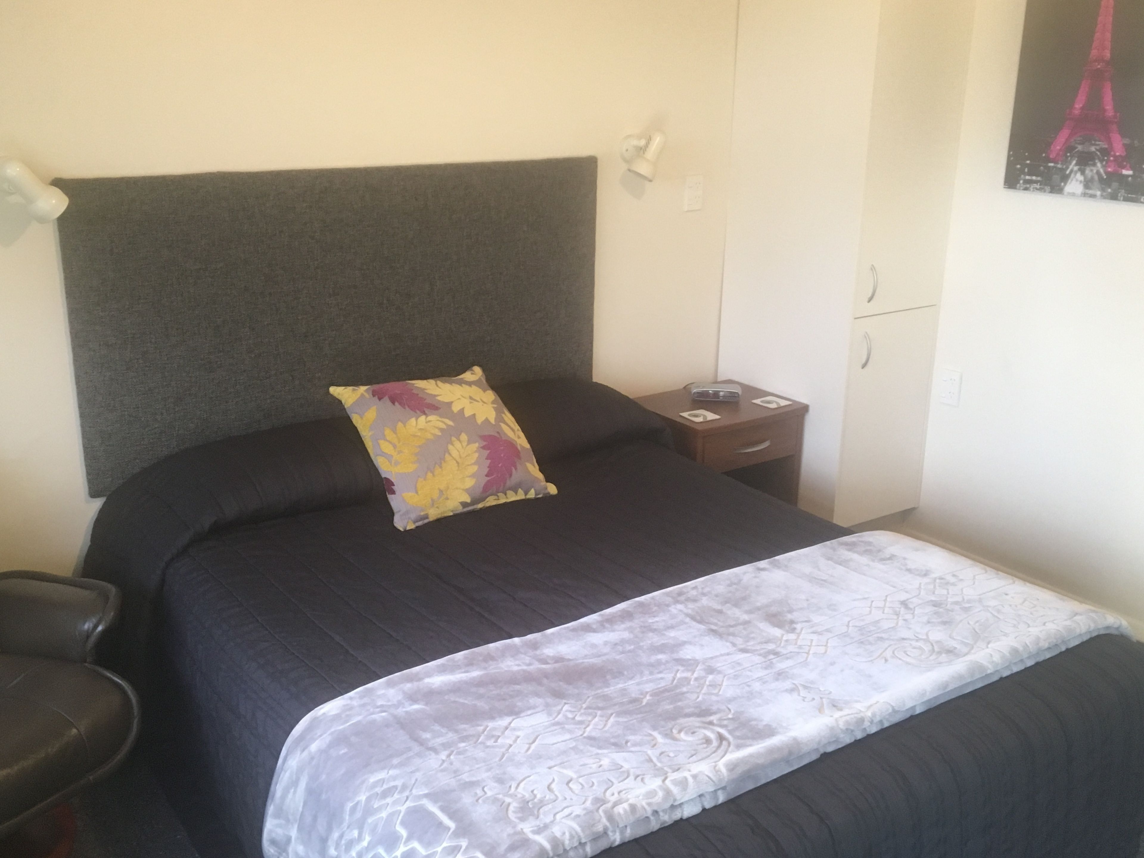 Una cama bien hecha con un cojín estampado, una mesita de noche con lámpara y un cuadro colgado en la pared.