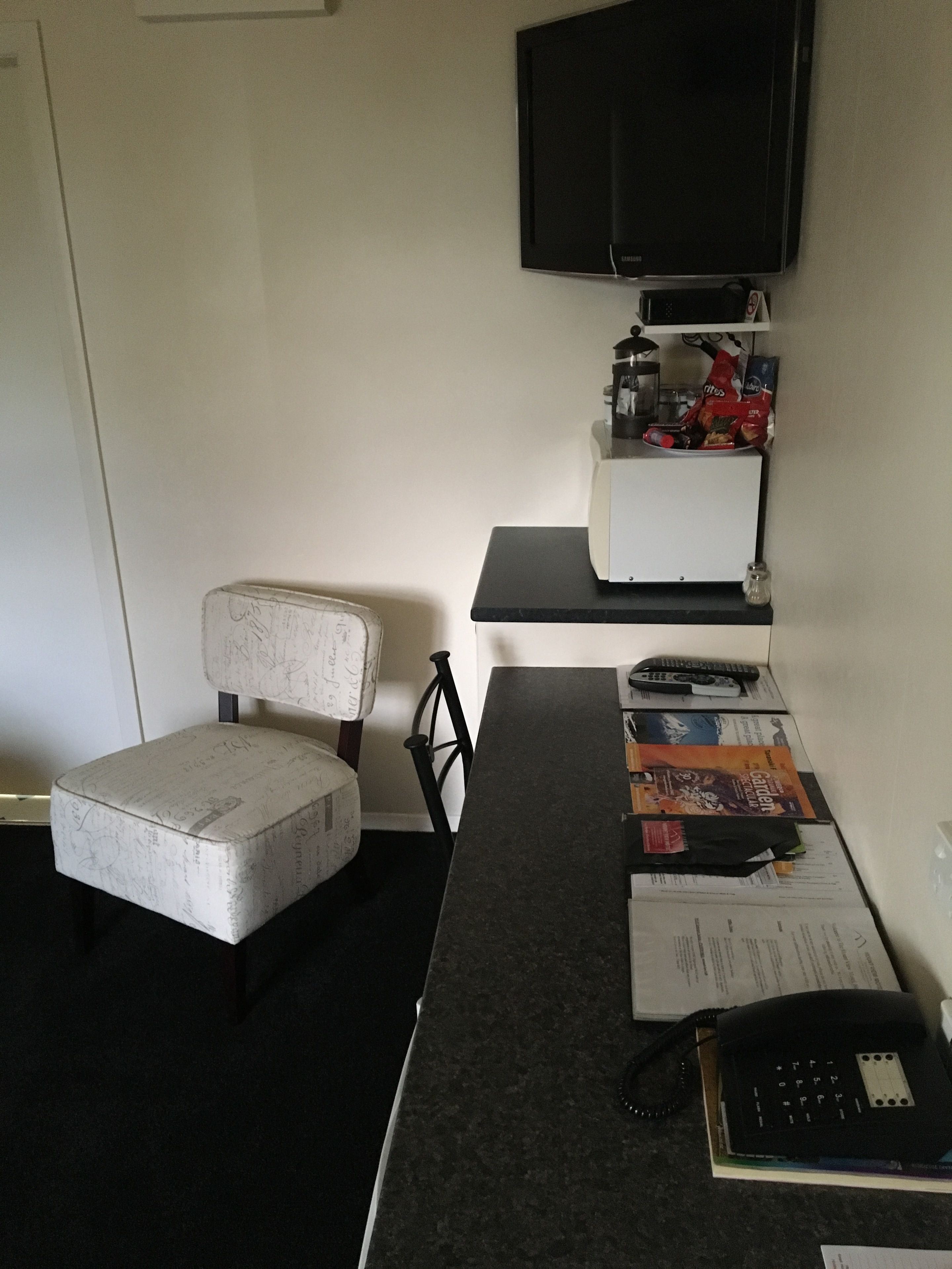 Une petite pièce avec un seul fauteuil blanc rembourré, un bureau noir avec un téléphone, une télécommande et quelques papiers. Un téléviseur à écran plat est fixé au mur, au-dessus du bureau.