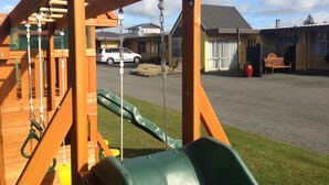 A playground with a swing set and slide, a parking area with a white car, and a building with a grey roof in the background.