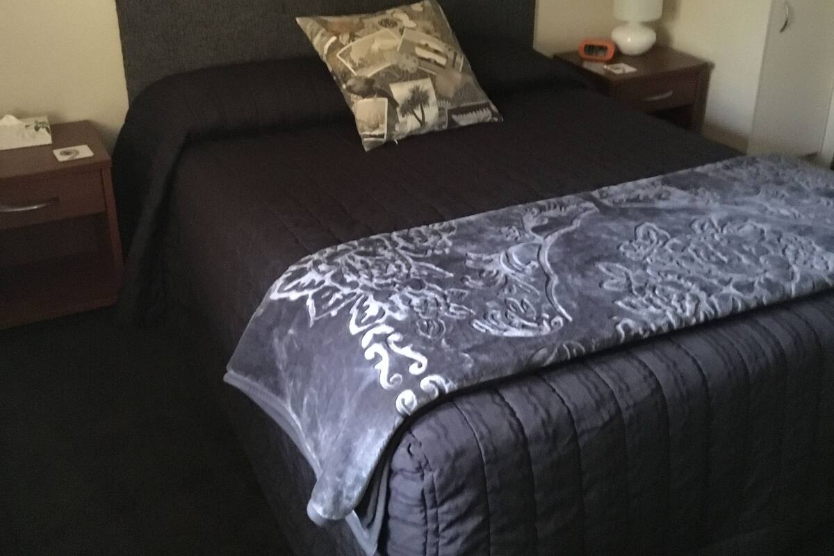 Una cama bien hecha con una colcha estampada, un almohadón con diseño de naturaleza y una mesita de noche con una lámpara y un reloj.