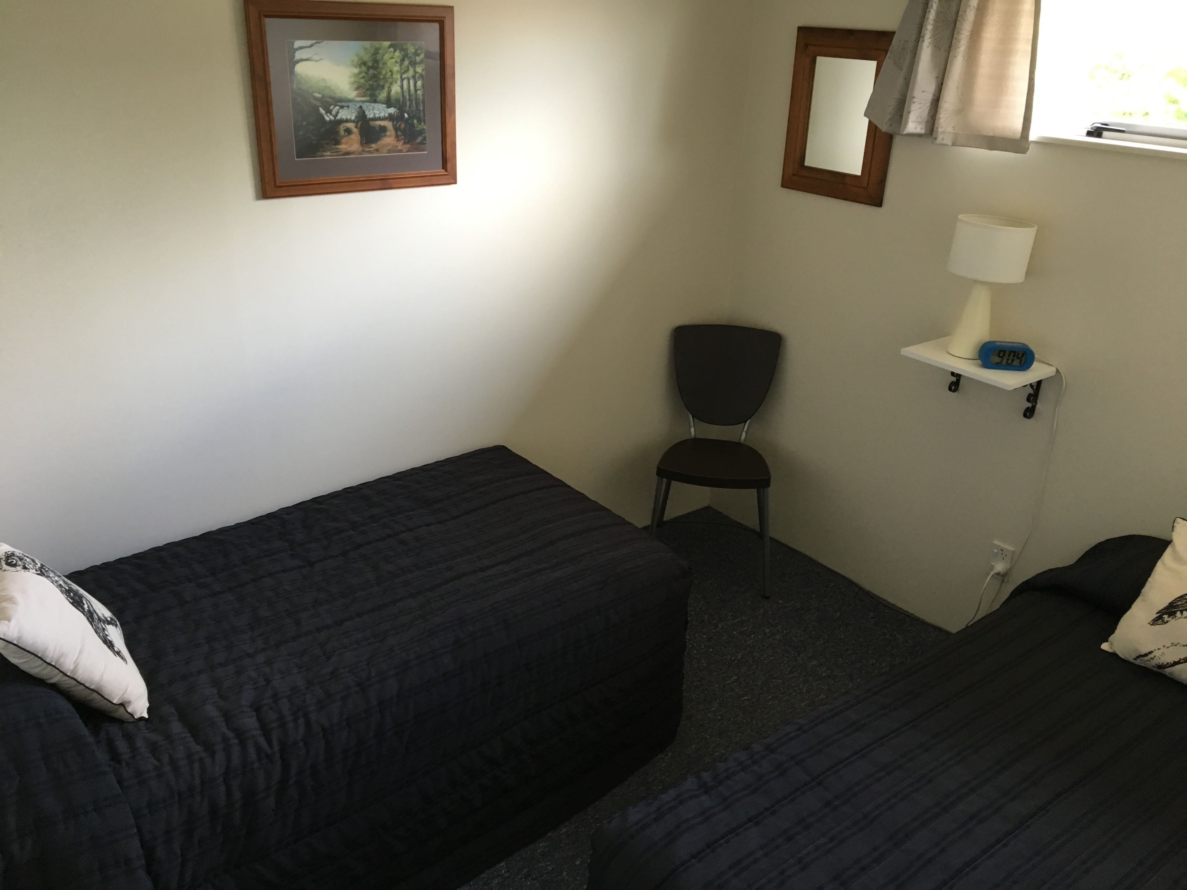 A small, dimly lit room with a single bed, a chair, a nightstand with a lamp, and a framed picture on the wall.