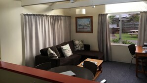 A room with a brown sofa, a wooden table, and a chair. There is a window with curtains, a framed picture on the wall, and a ceiling light fixture.