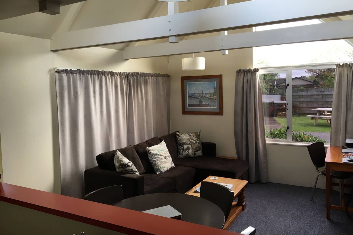 Una habitación con un sofá marrón, una mesa de madera y una silla. Se ve una ventana con cortinas, un cuadro enmarcado en la pared y una lámpara en el techo.