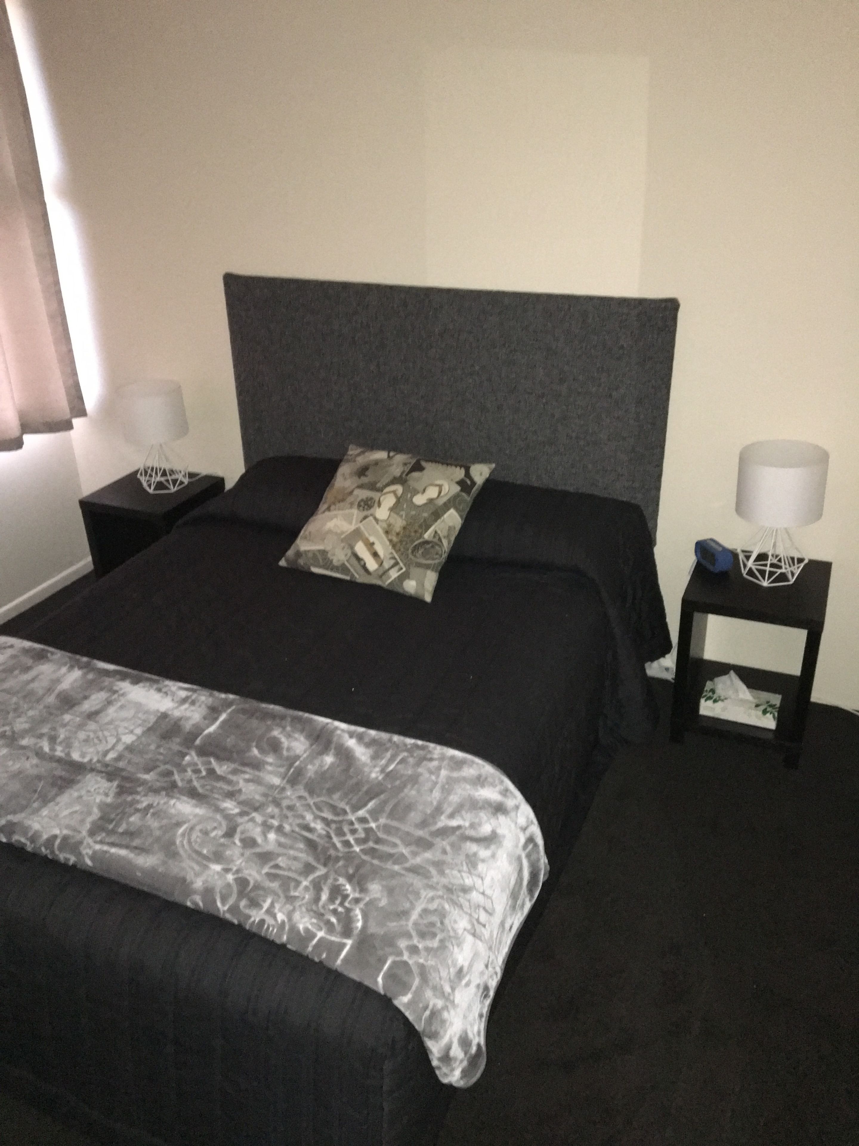 Ein ordentlich bezogenes Bett mit grauem Kopfteil, einem gemusterten Kissen und einer grauen Decke. Es gibt zwei Nachttische, auf einem steht eine Lampe und eine kleine Pflanze.