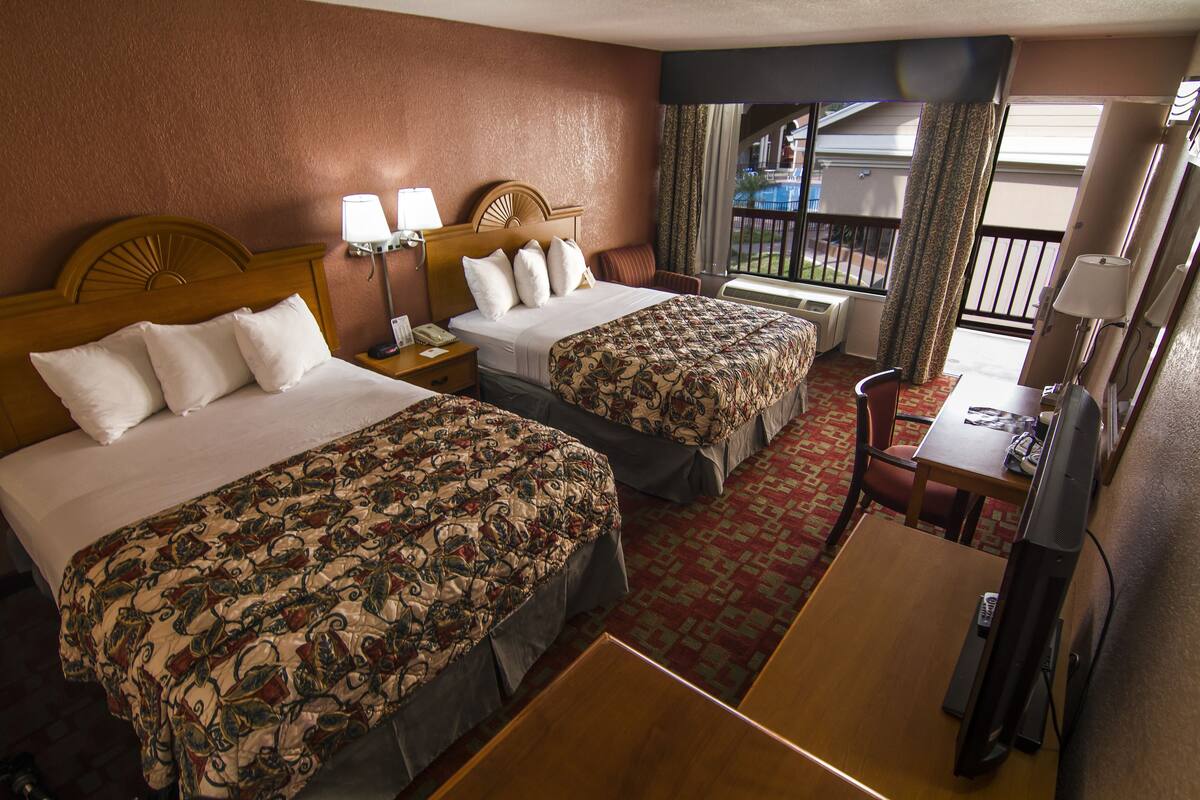 A hotel room with two beds, a desk, a chair, a TV, and a window with a view of a building and a balcony.