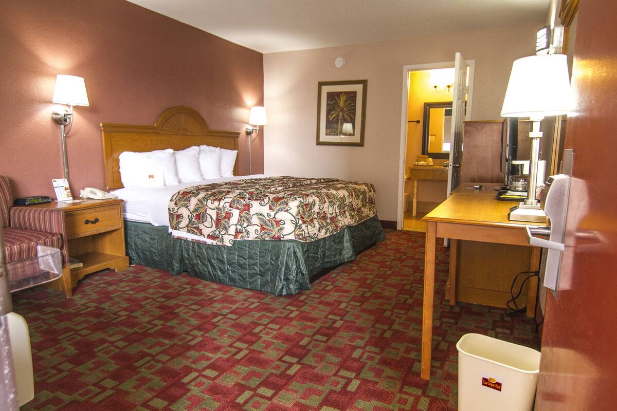 A hotel room with a bed, a desk, a chair, a nightstand, a lamp, and a trash bin.