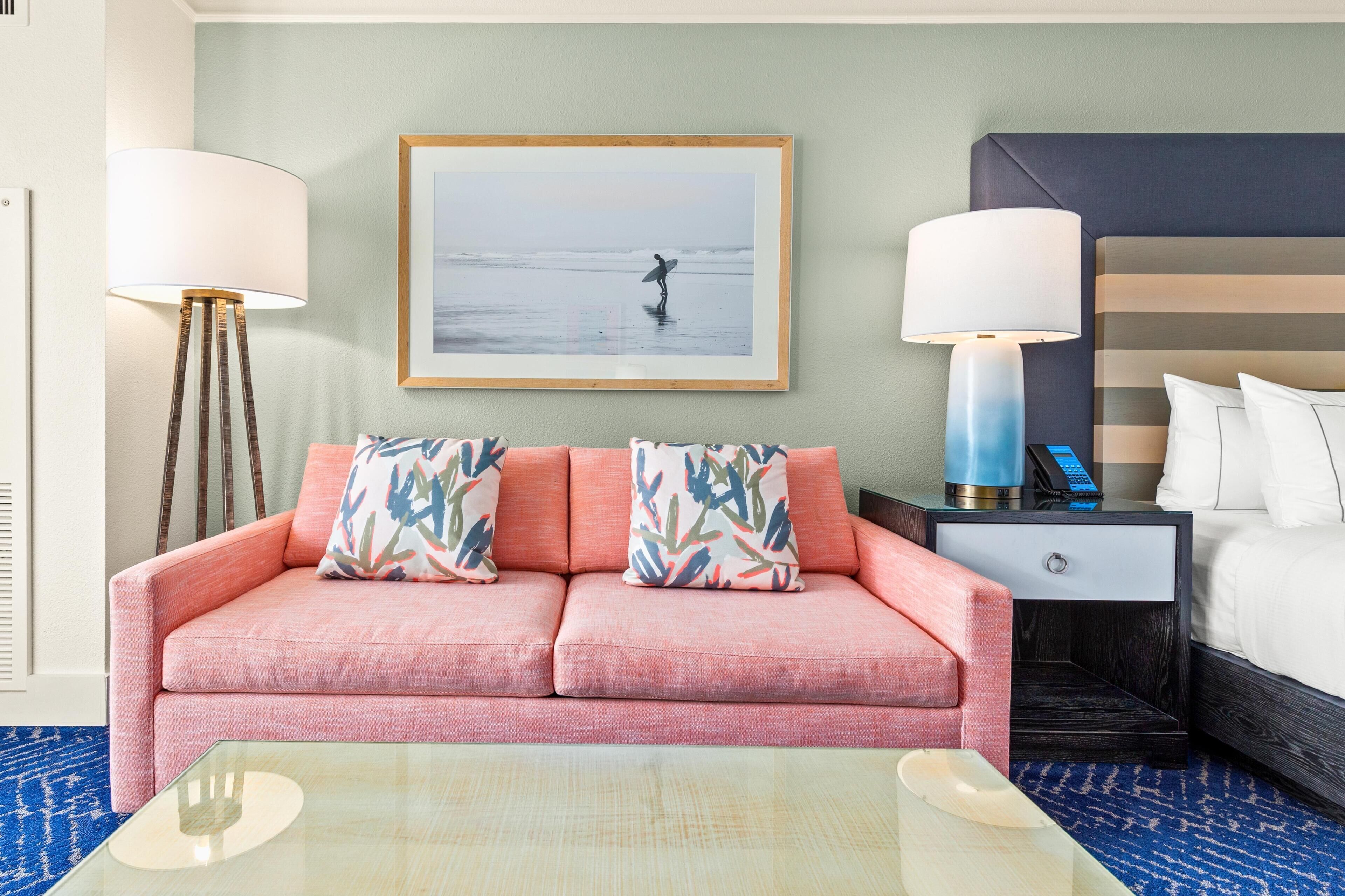 A hotel room with a pink sofa, a framed picture of a surfer, a bedside table with a lamp, and a bed with white linens.