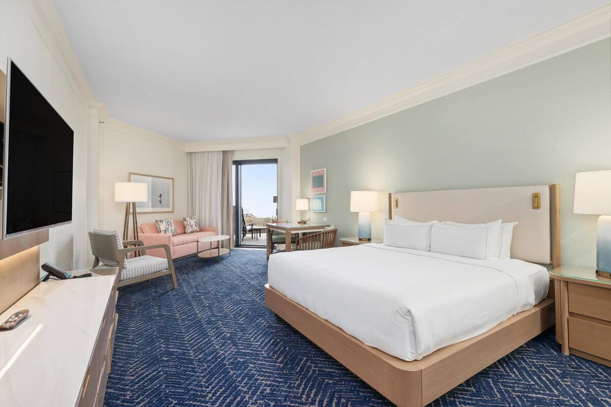 A hotel room with a large bed, a desk with a TV, a sofa, a dining table, and a view of the outdoors.