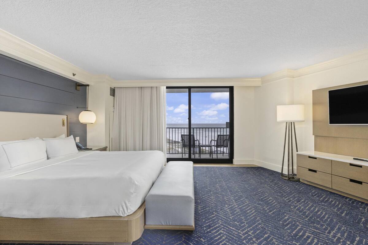 A hotel room with a bed, a bench, a TV, a desk, and a balcony with a view.