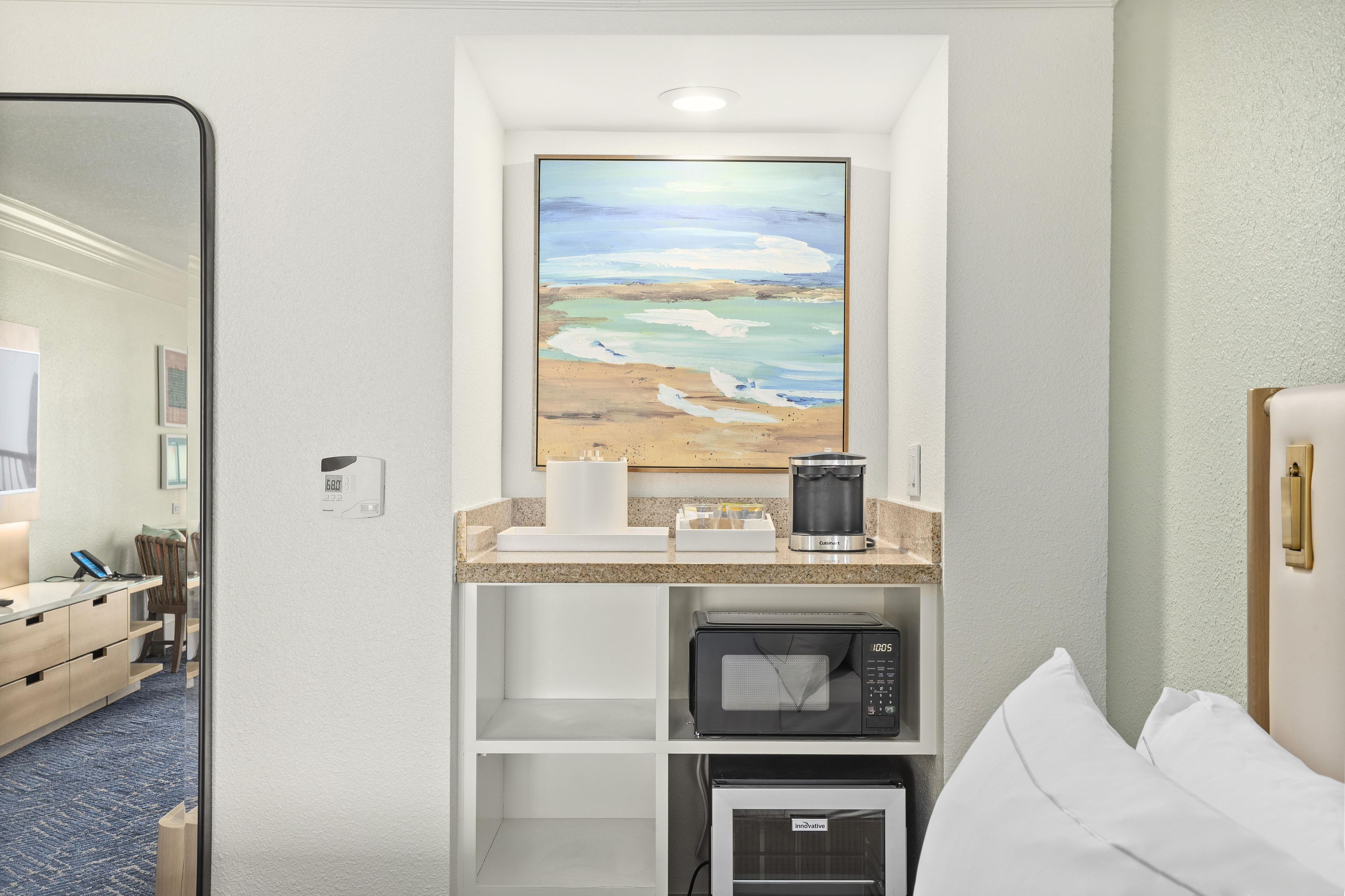 Habitación de hotel con un cuadro de una escena de playa, una kitchenette con cafetera, microondas y una cama con ropa blanca.