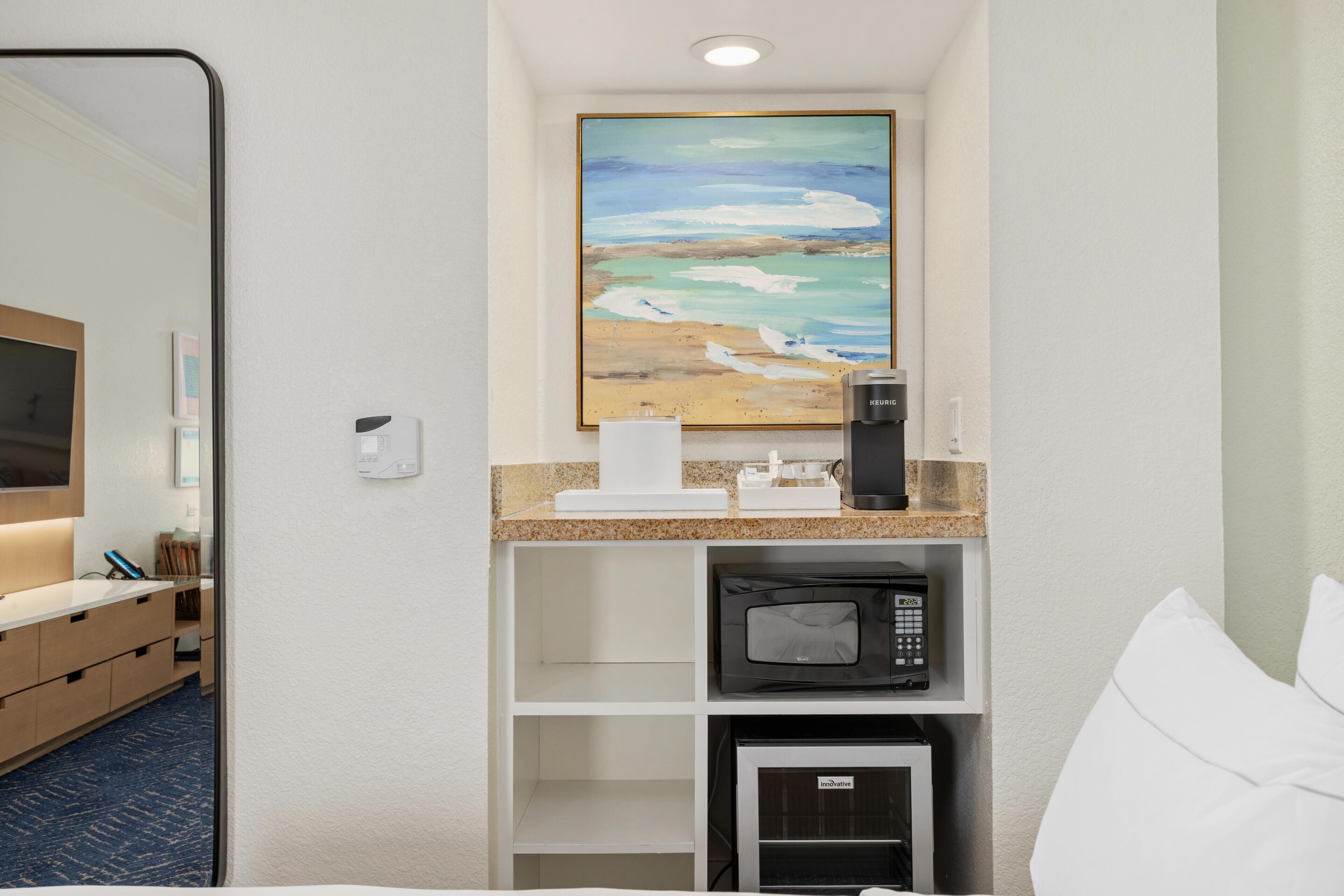 Ein Hotelzimmer mit Bett, Mikrowelle, Kaffeemaschine, einem Bild mit Strandmotiv und einem kleinen Kühlschrank.
