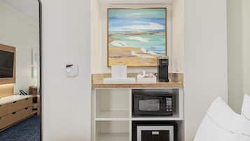 A hotel room with a bed, a microwave, a coffee maker, a painting of a beach scene, and a small refrigerator.