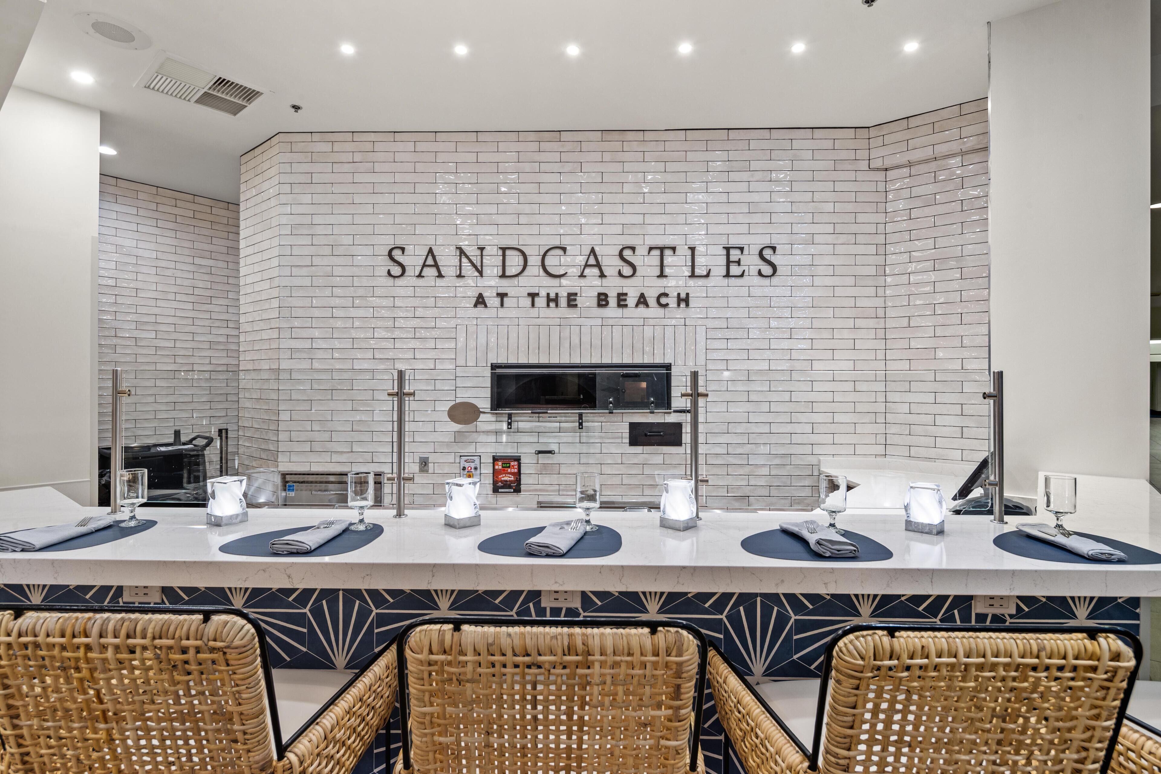Un restaurant à thème balnéaire, avec un long comptoir, des chaises en osier et un mur recouvert de carreaux sur lequel est inscrit le nom « SANDCASTLES AT THE BEACH ».