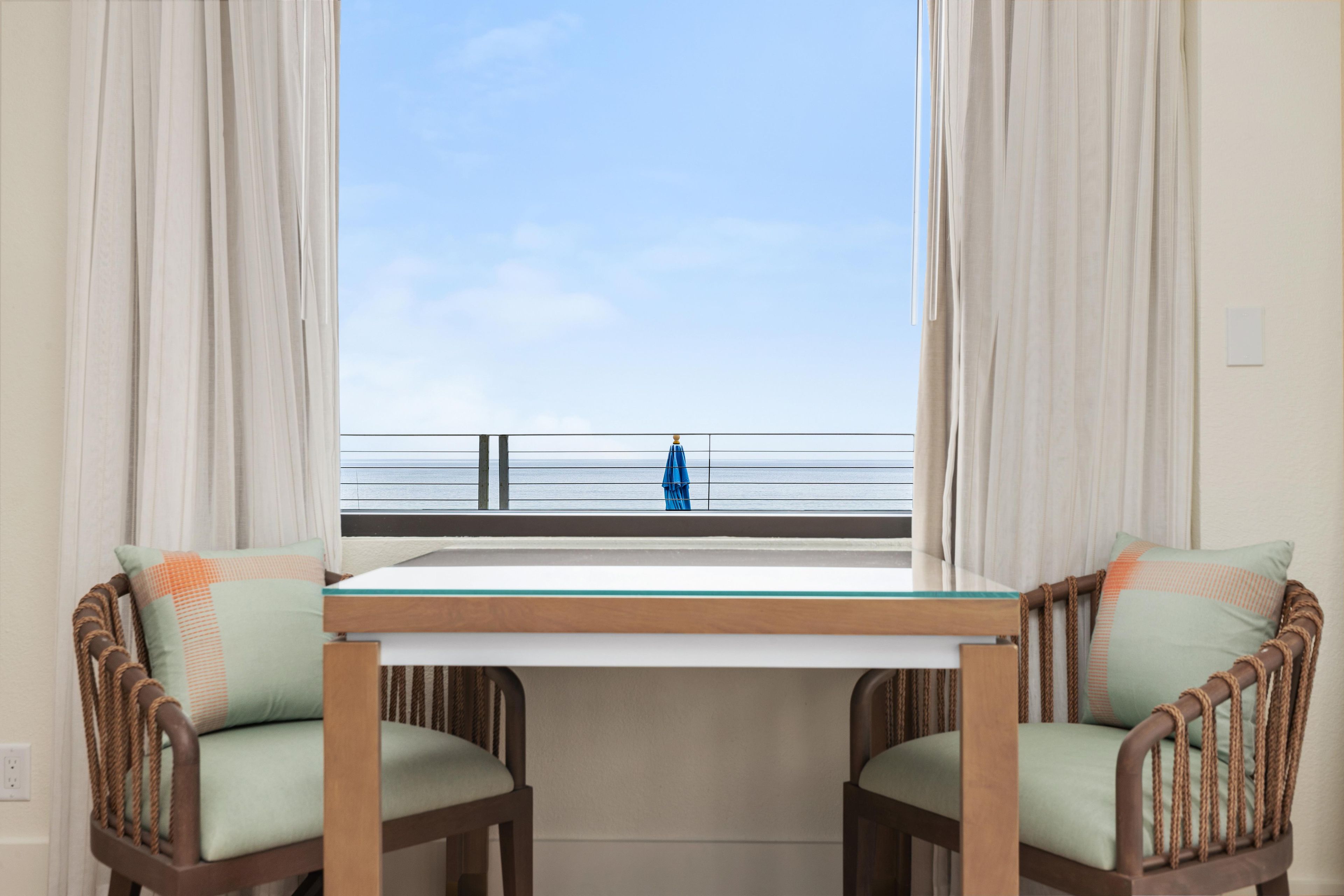 Een kamer met uitzicht op zee, met een tafel, stoelen, een raam en gordijnen.