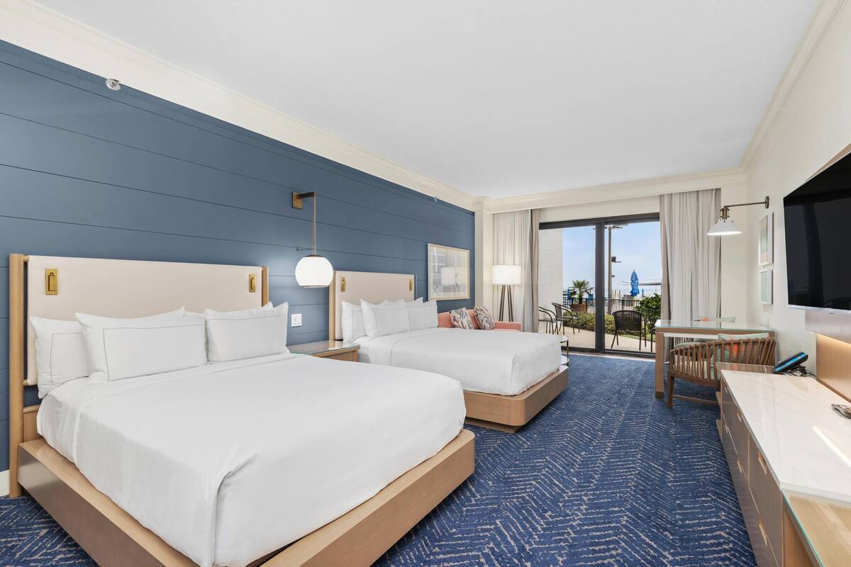 A hotel room with two beds, a desk, a TV, and a balcony with outdoor seating.