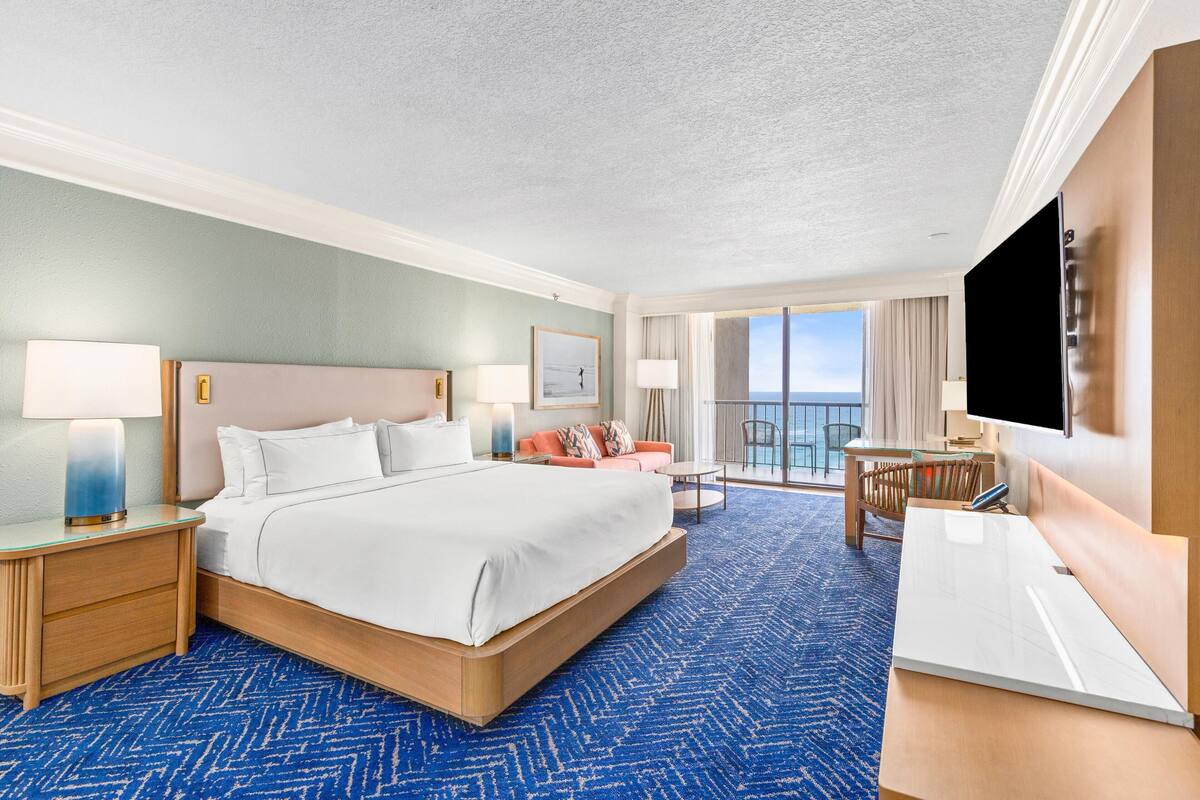 A hotel room with a large bed, a desk, a TV, and a balcony with a view.