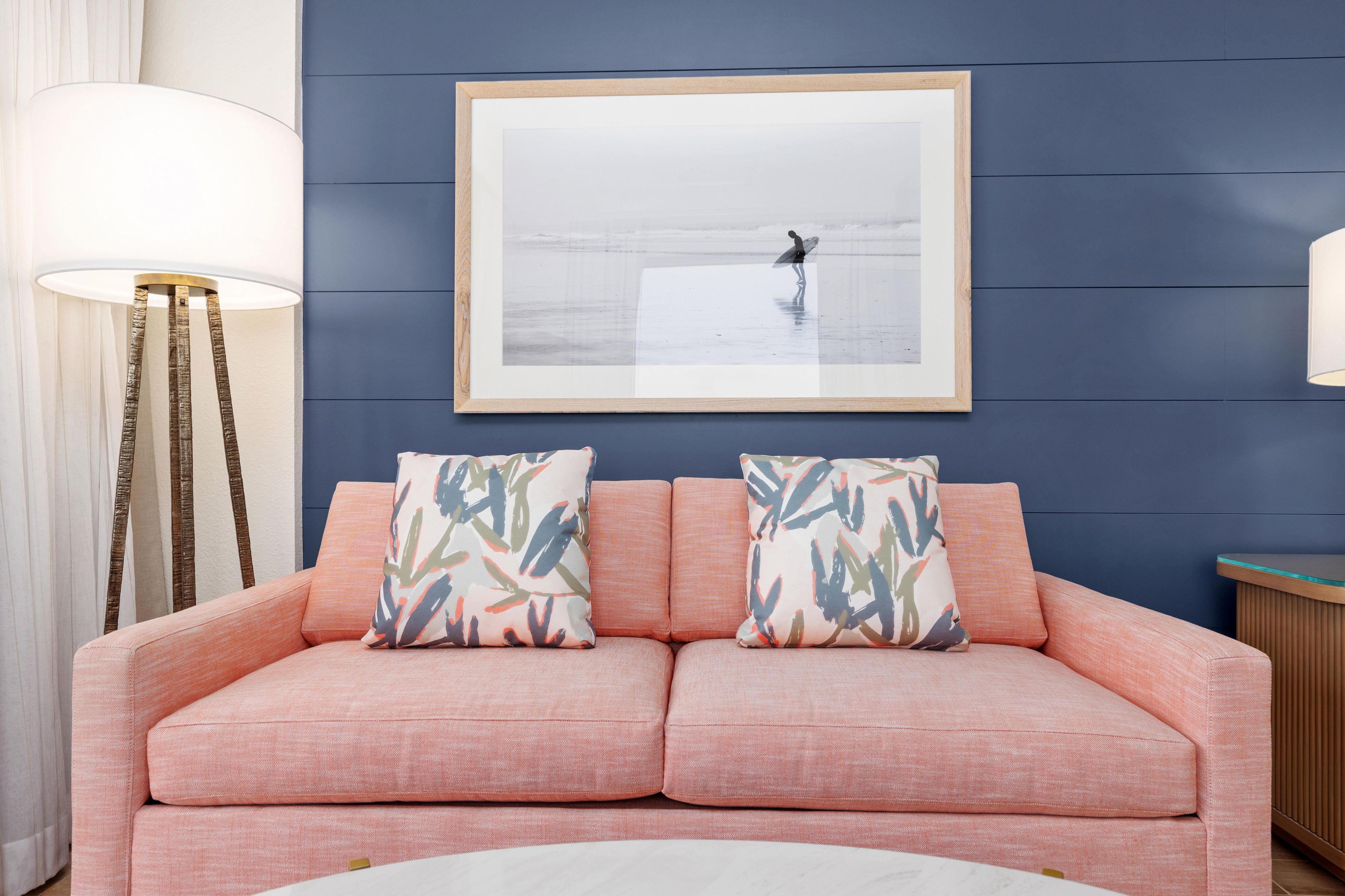 Un canapé rose agrémenté de deux coussins à motifs, une photographie encadrée au-dessus, et une lampe sur pied à gauche.