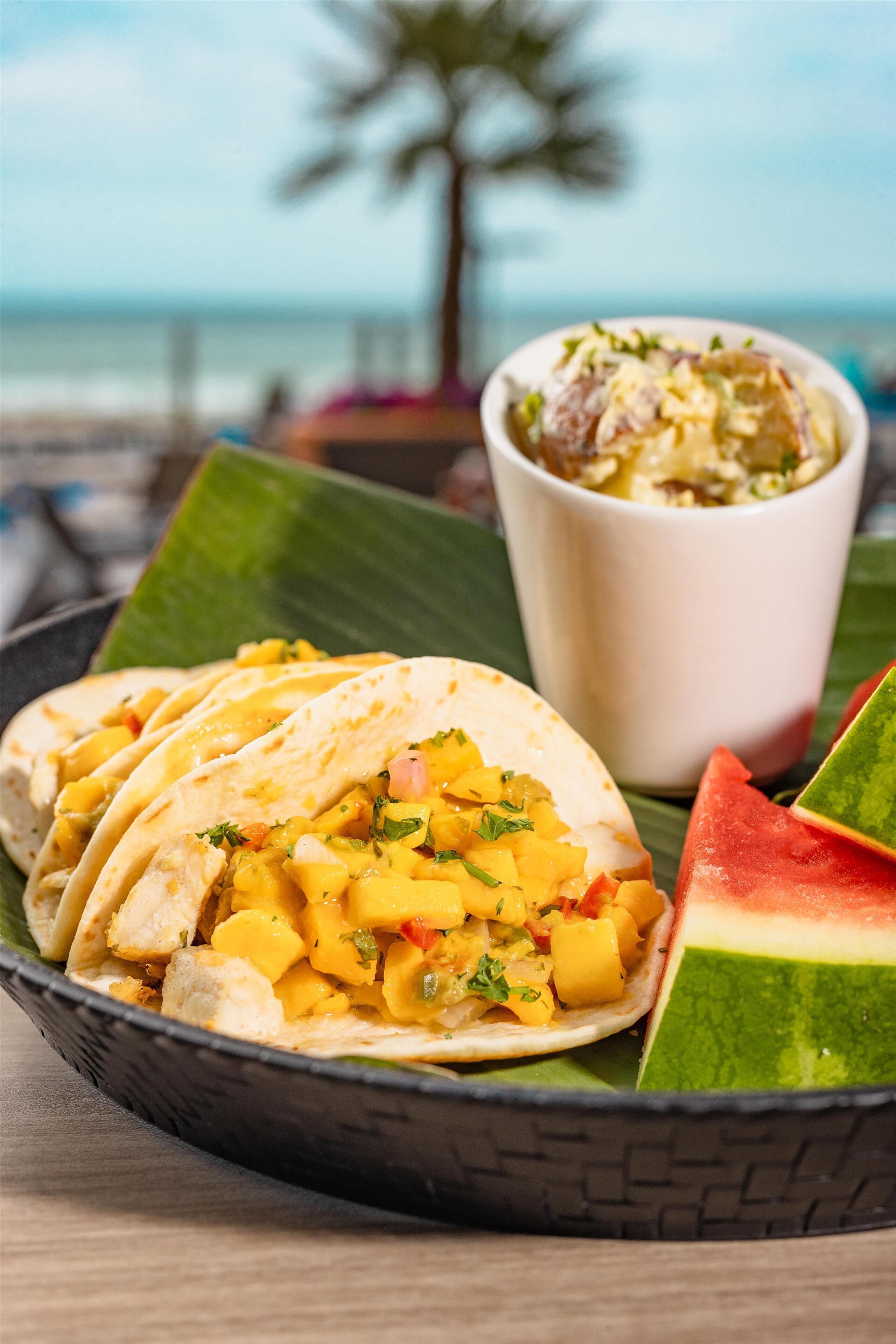 A plate of tacos with mango and a bowl of salad on a table with a view of the ocean.