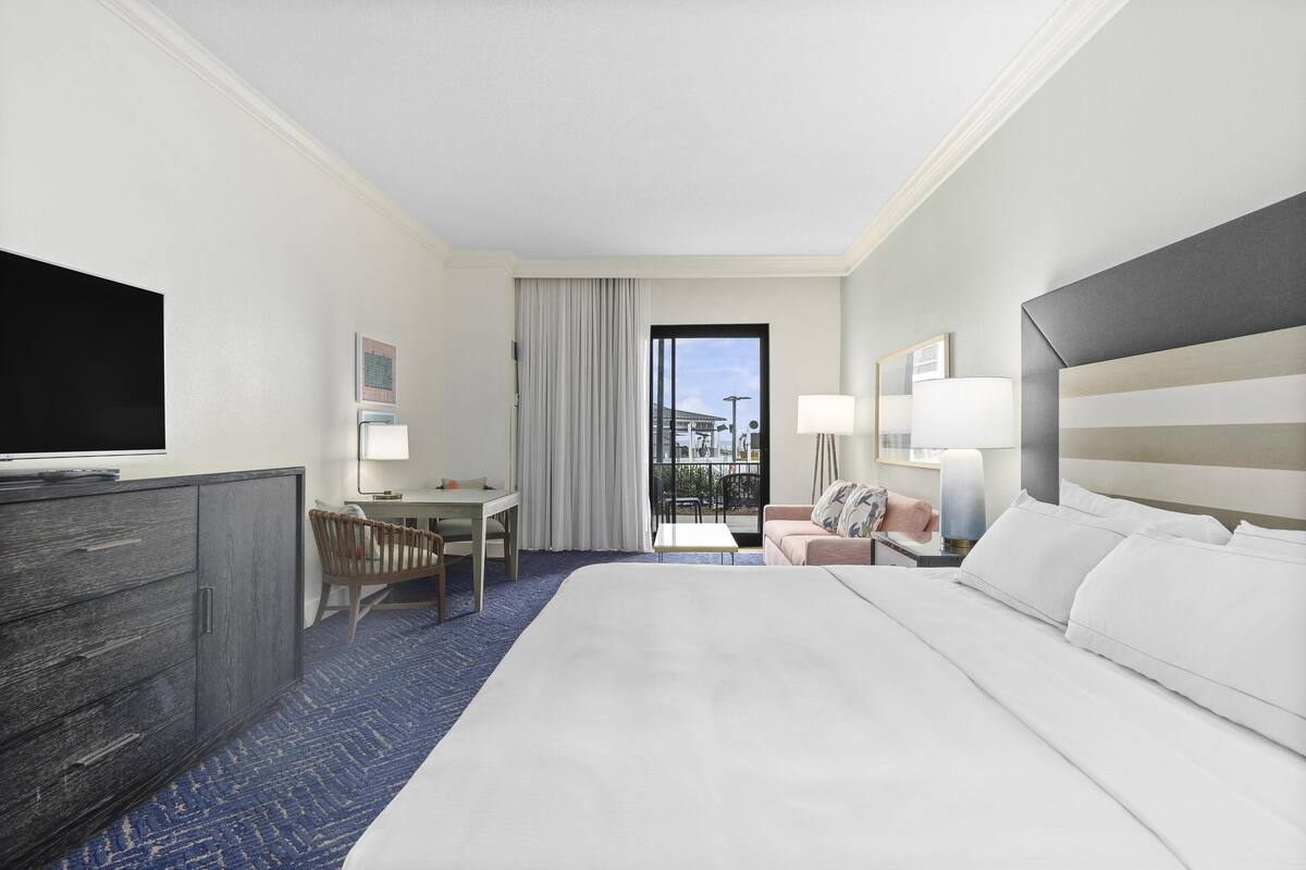 A hotel room with a large bed, a desk, a chair, a television, and a balcony with a view.