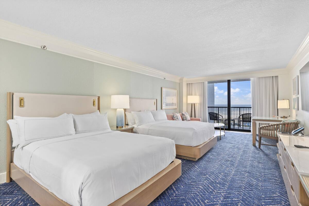 A hotel room with two beds, a desk, a chair, and a balcony with a view.