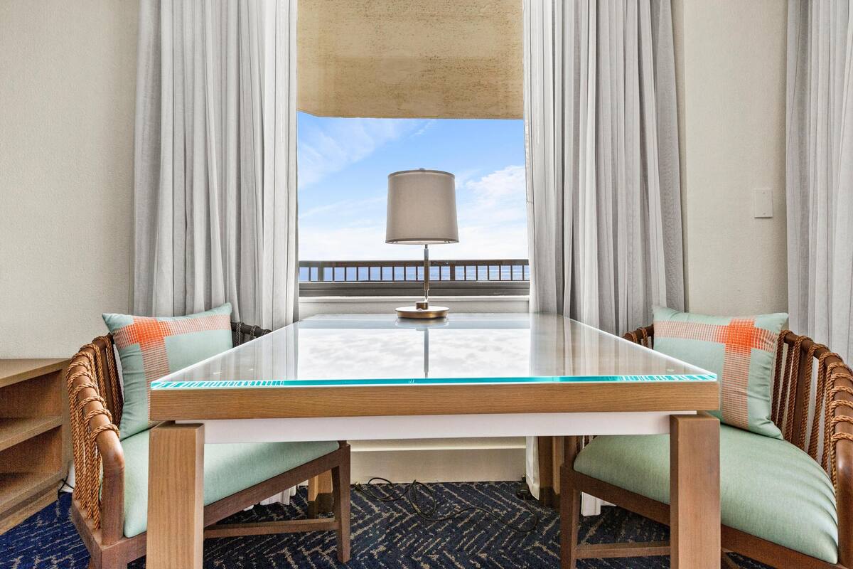 A room with a glass-top table, two cushioned chairs, a lamp, and a view of the sea through curtains.