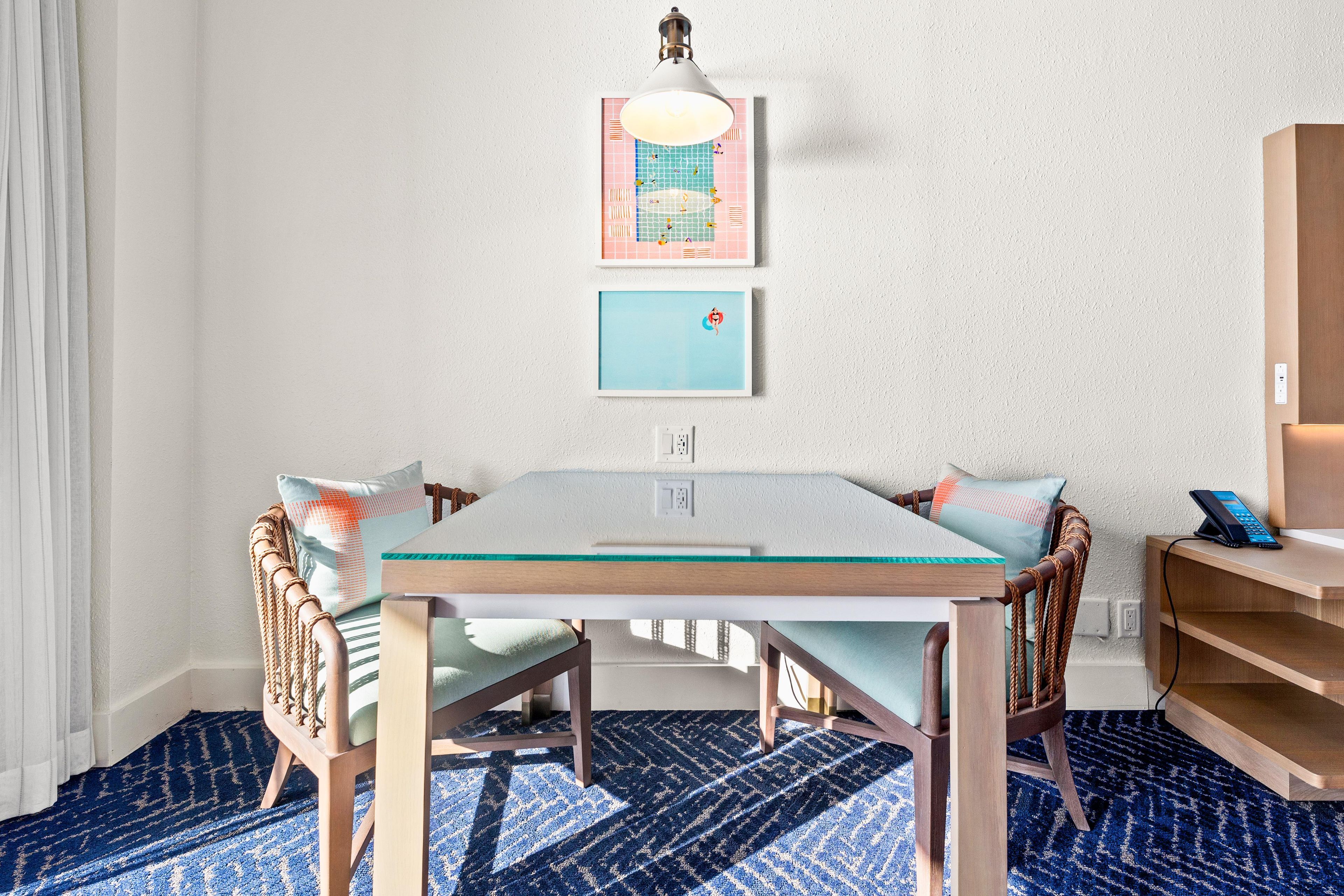 Un área de comedor con una mesa de vidrio, cuatro sillas con respaldo tejido, una lámpara fijada a la pared y dos obras de arte enmarcadas.
