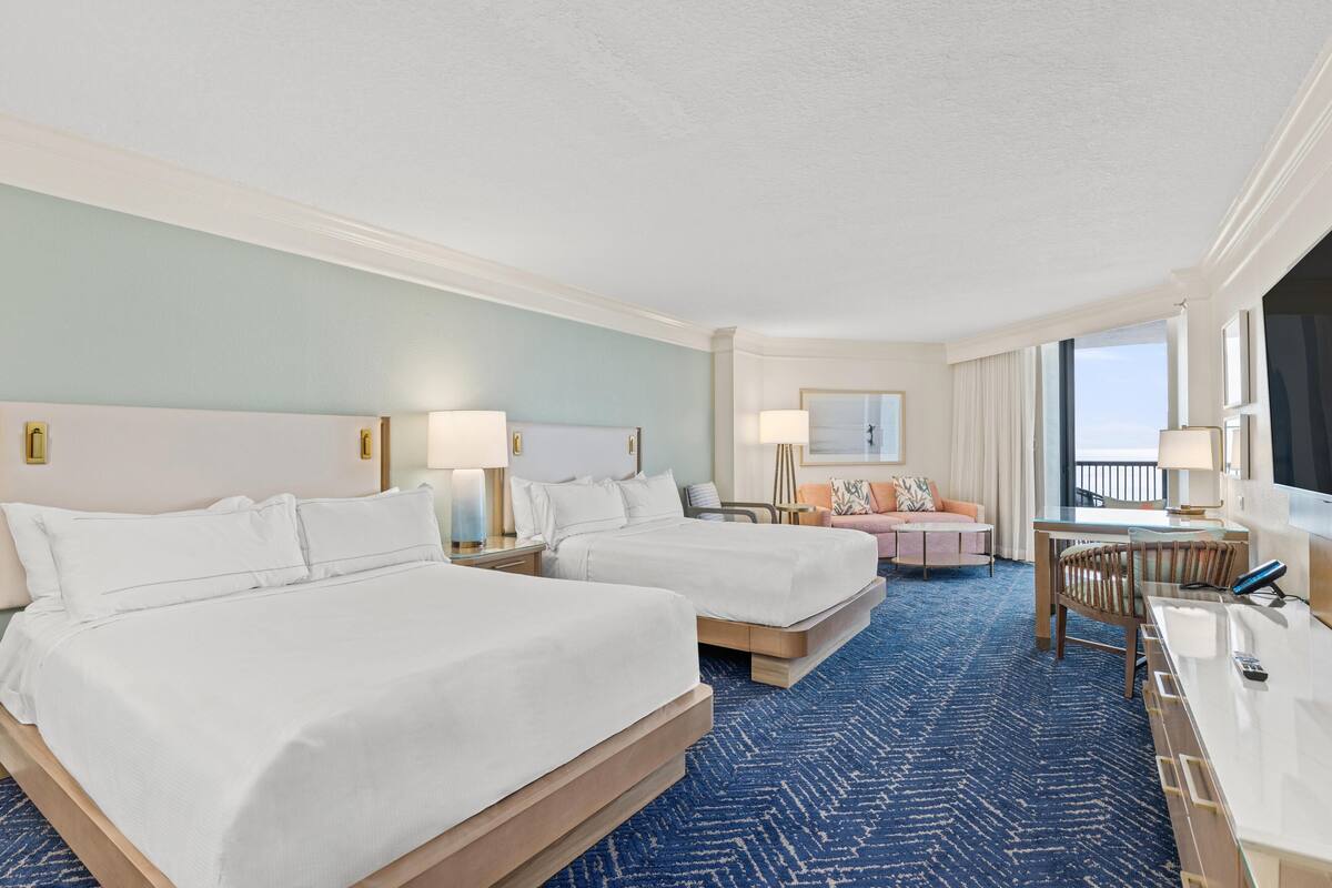 A hotel room with two beds, a desk, a chair, a sofa, and a large window.