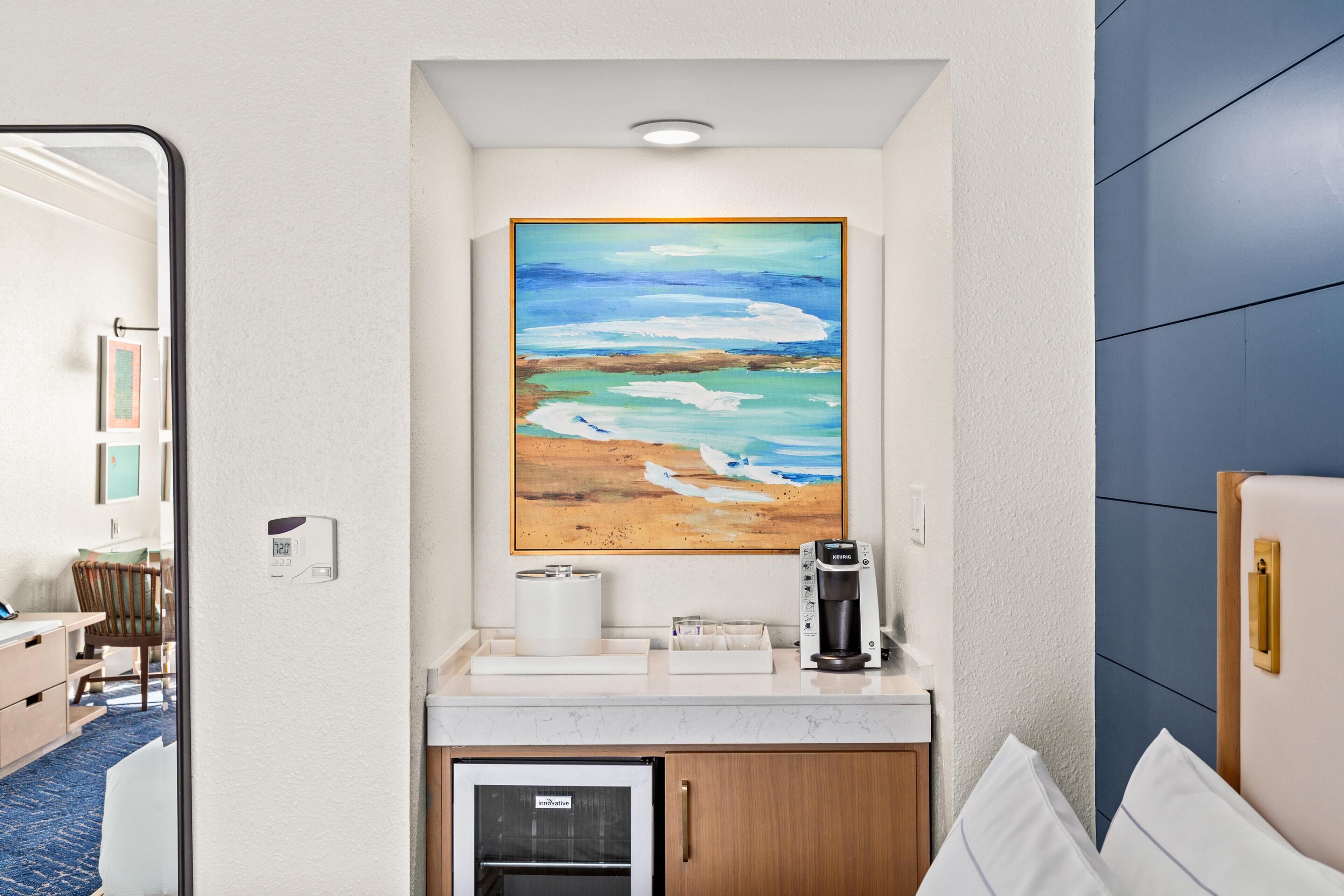 A hotel room with a kitchenette, a painting of a beach scene, a small refrigerator, and a bedside table.