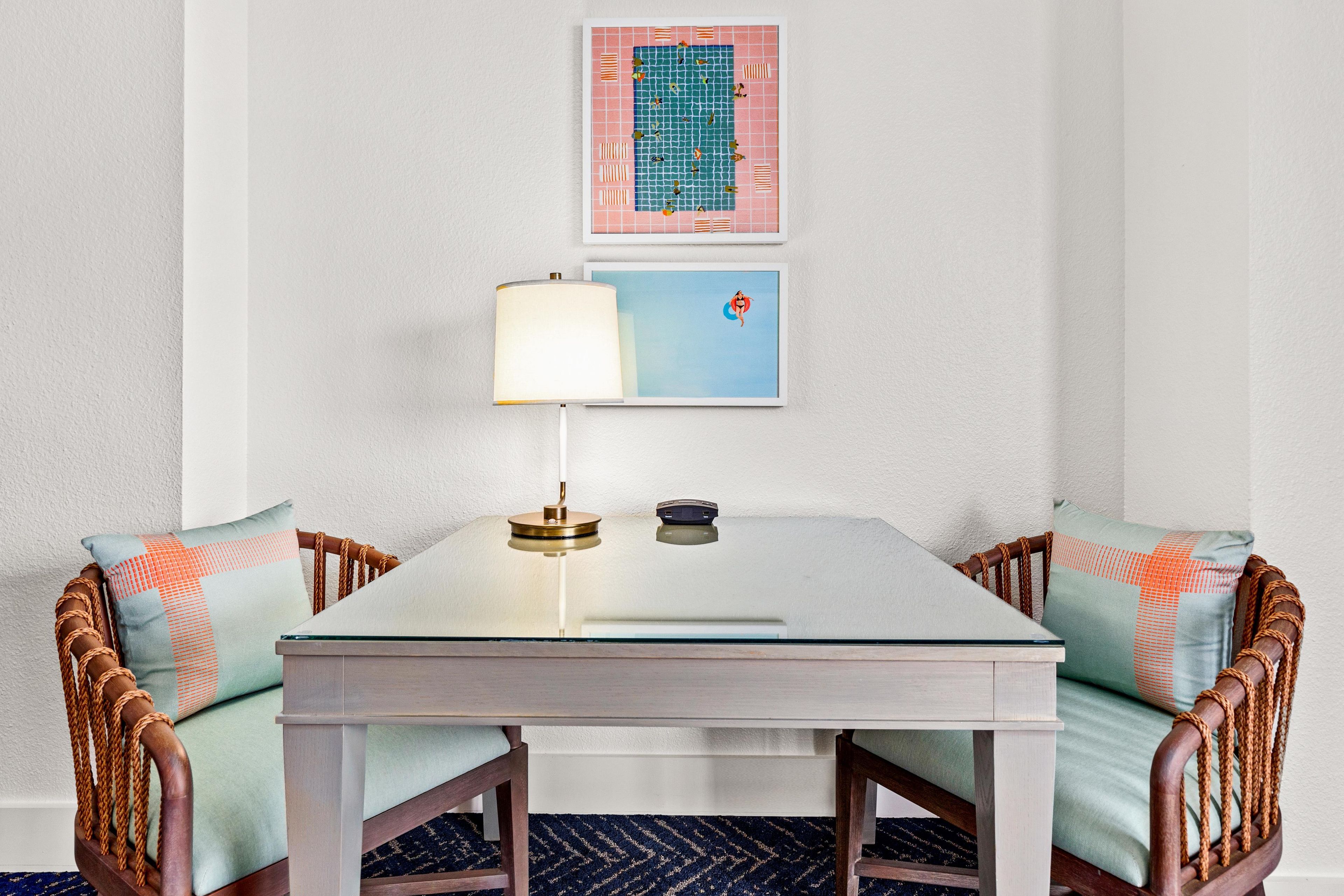 Een kamer met een witte tafel, twee rieten stoelen, een lamp en twee foto’s in lijsten aan de muur.