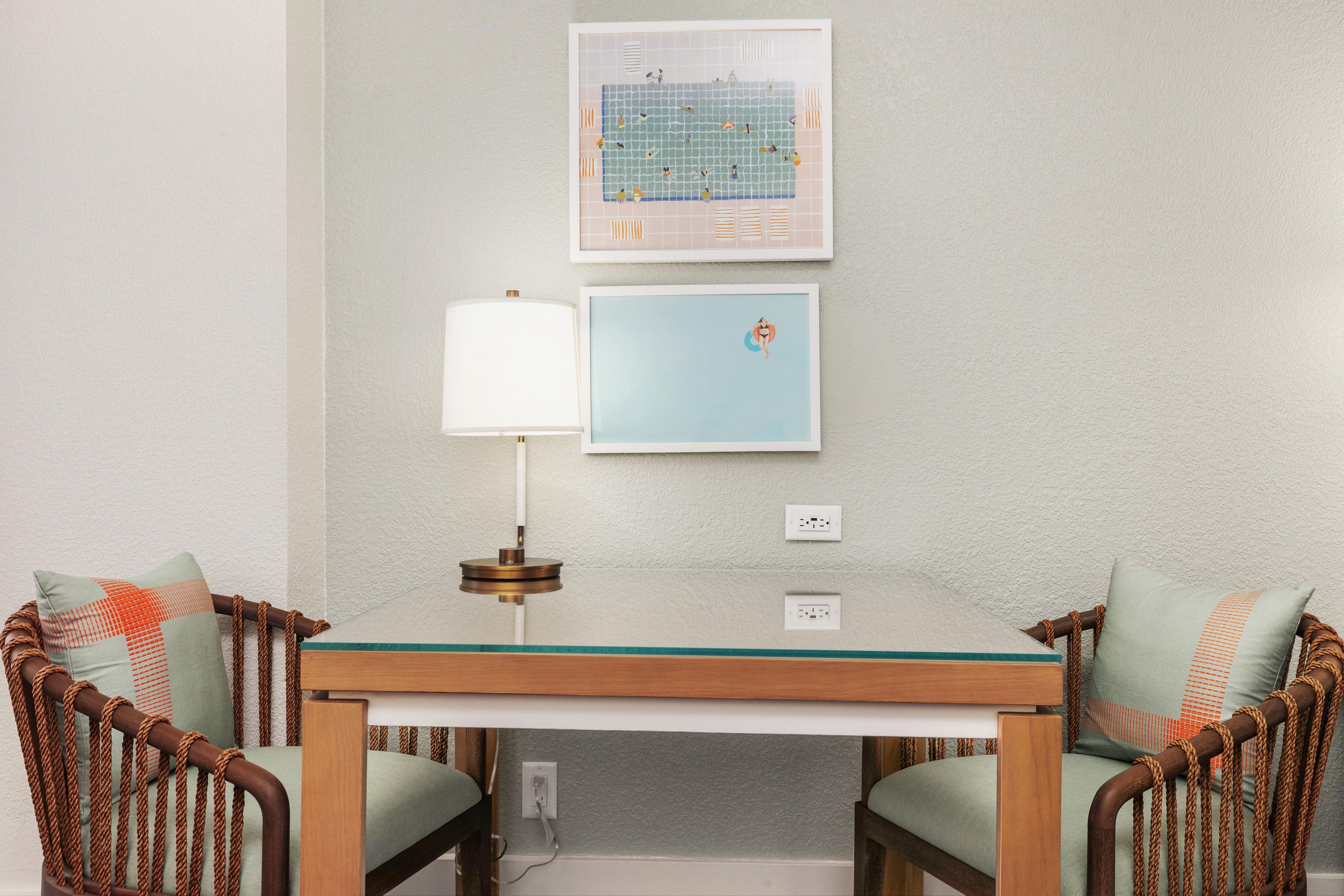 A room with a glass-top table, two wicker chairs, a wall-mounted lamp, and two framed pictures.