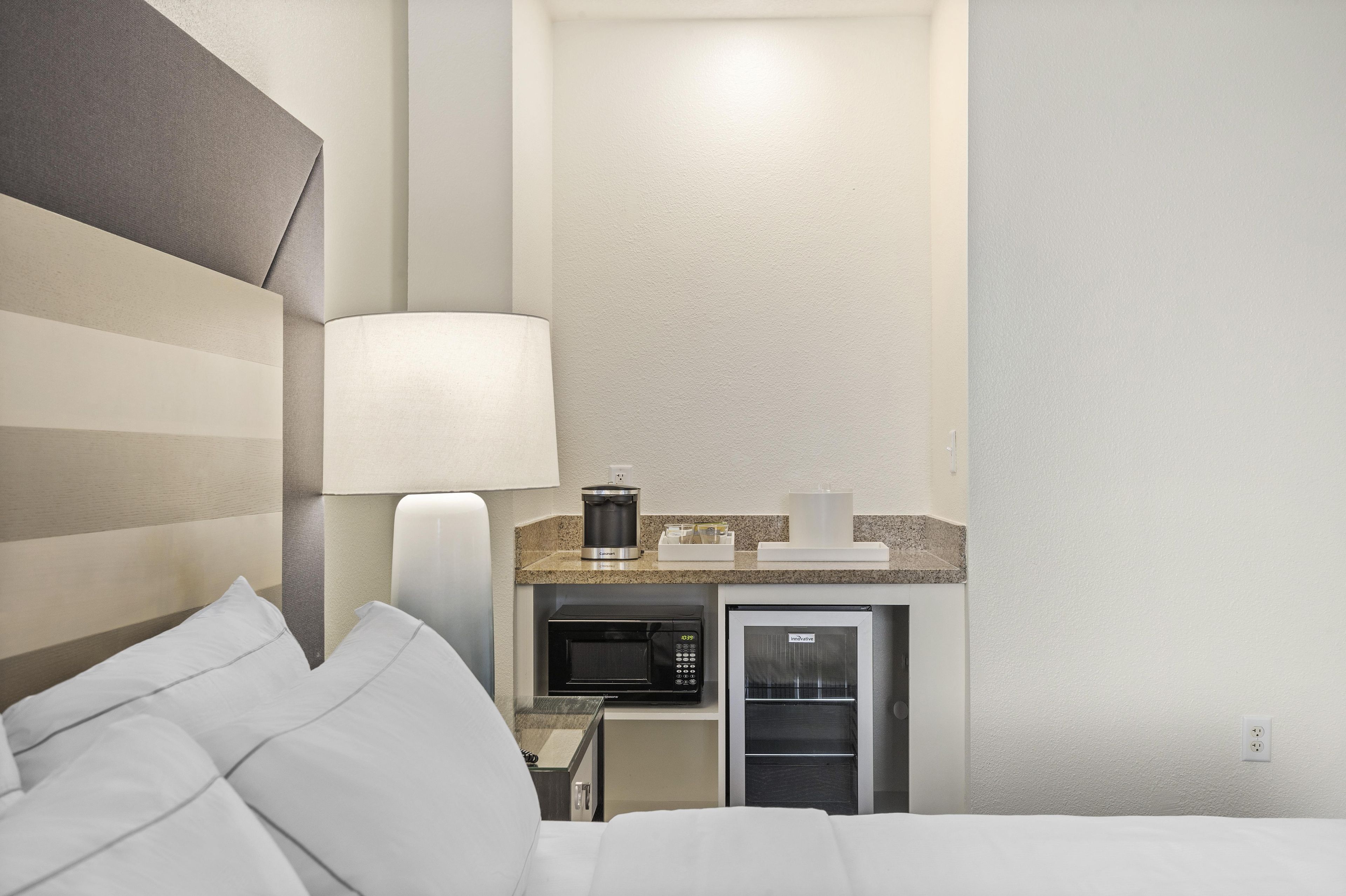 A hotel room with a bed, a bedside lamp, a microwave, and a refrigerator.