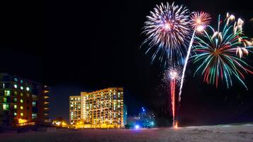 A hotel with multiple floors and balconies, a beach in the foreground, and fireworks in the background.