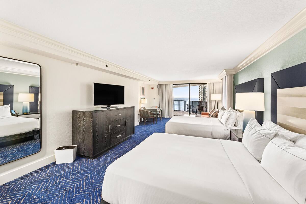 A hotel room with two beds, a TV, a desk, and a balcony with a view.