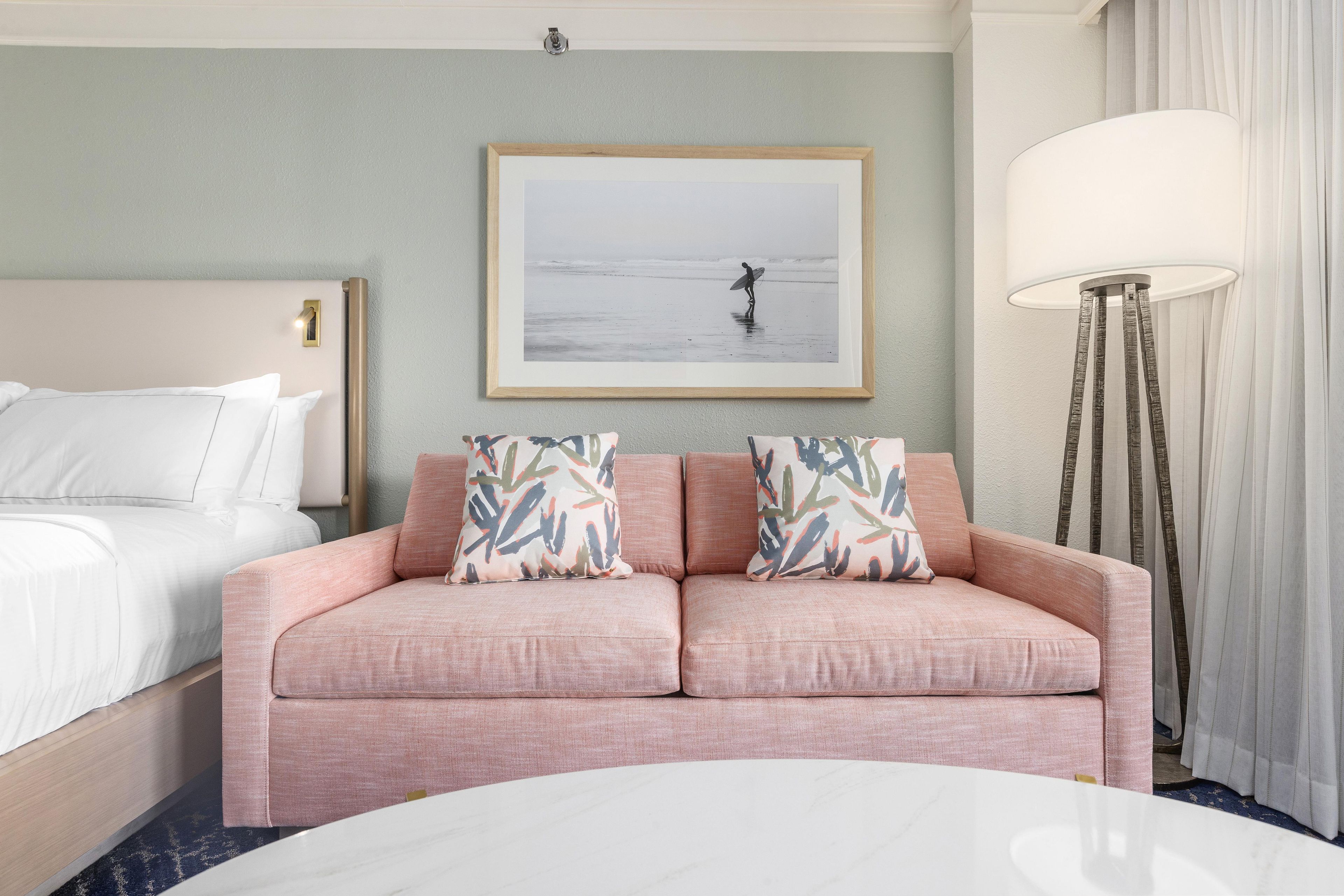 Ein Hotelzimmer mit einem Bett, einem rosa Sofa mit gemusterten Kissen, einem gerahmten Bild von einer Strandansicht und einer Stehlampe.