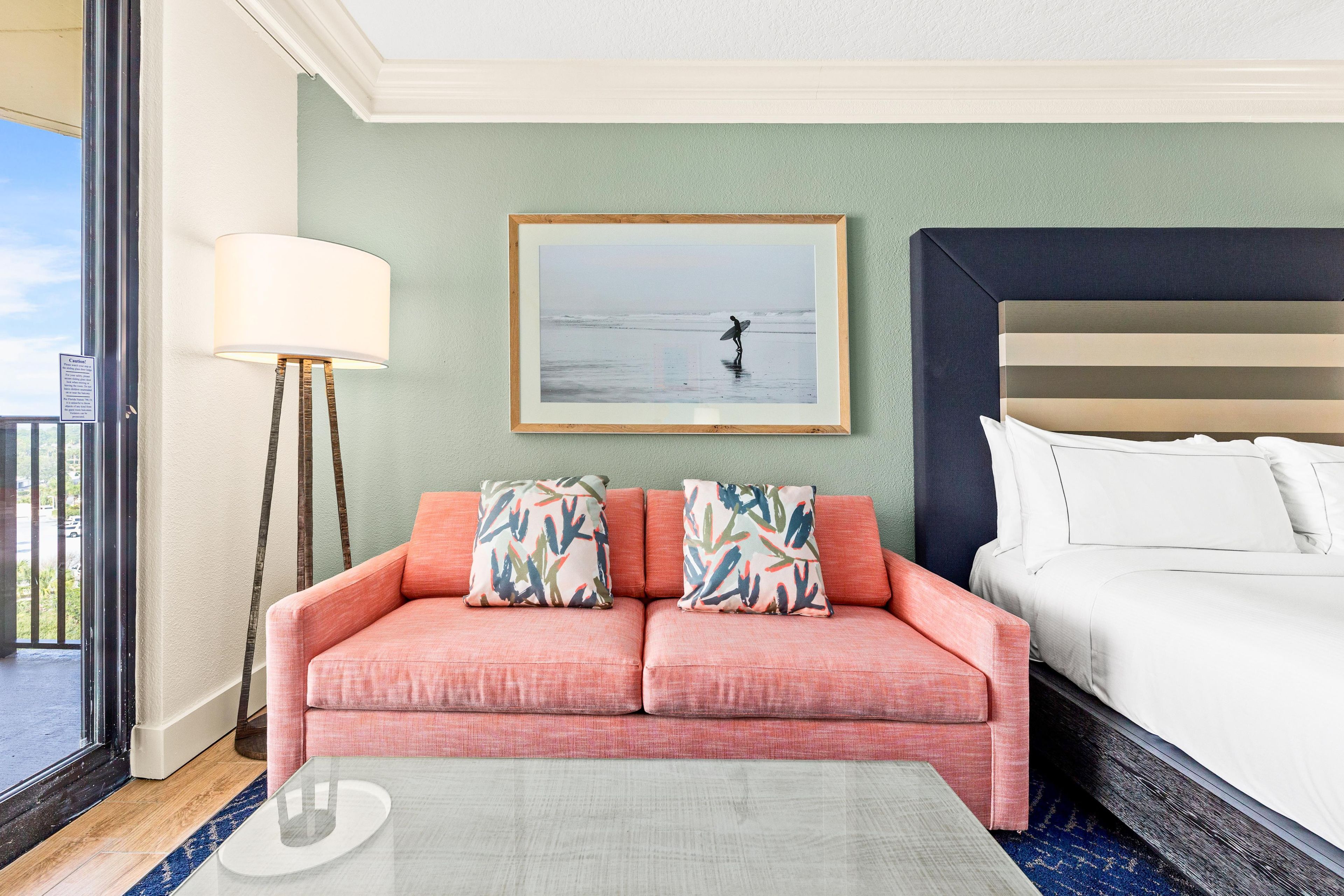 Ein Hotelzimmer mit einer Couch, einem Bett, einem gerahmten Bild mit Strandmotiv und einer Stehlampe.