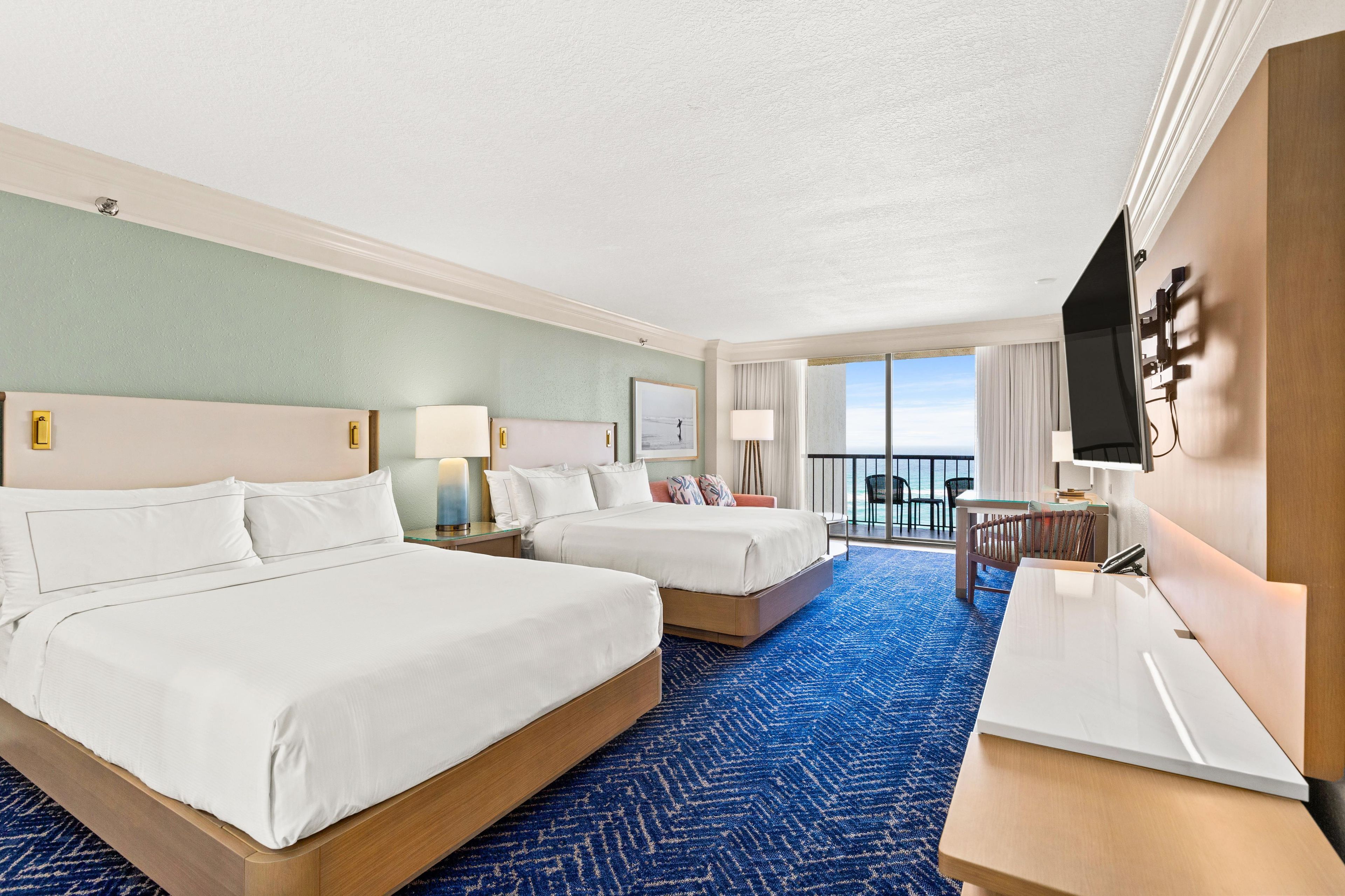 Habitación de hotel con dos camas, un escritorio, un televisor y un balcón con vistas.