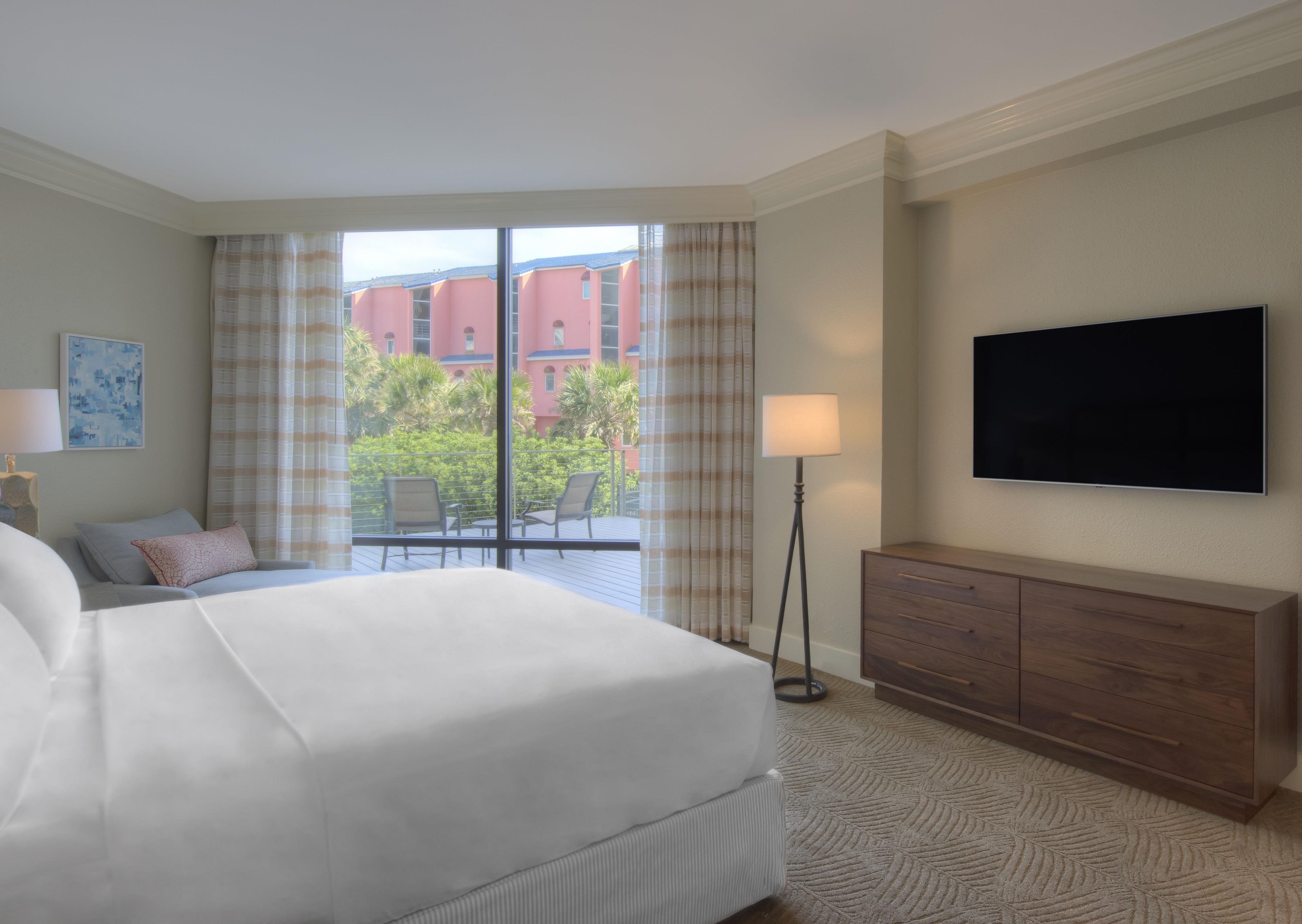 A hotel room with a bed, a flat-screen TV, a desk with a lamp, a view of buildings and greenery outside.