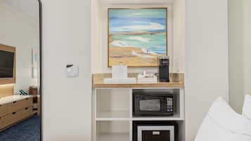A hotel room with a kitchenette area, a microwave, a coffee maker, and a painting of a beach scene.