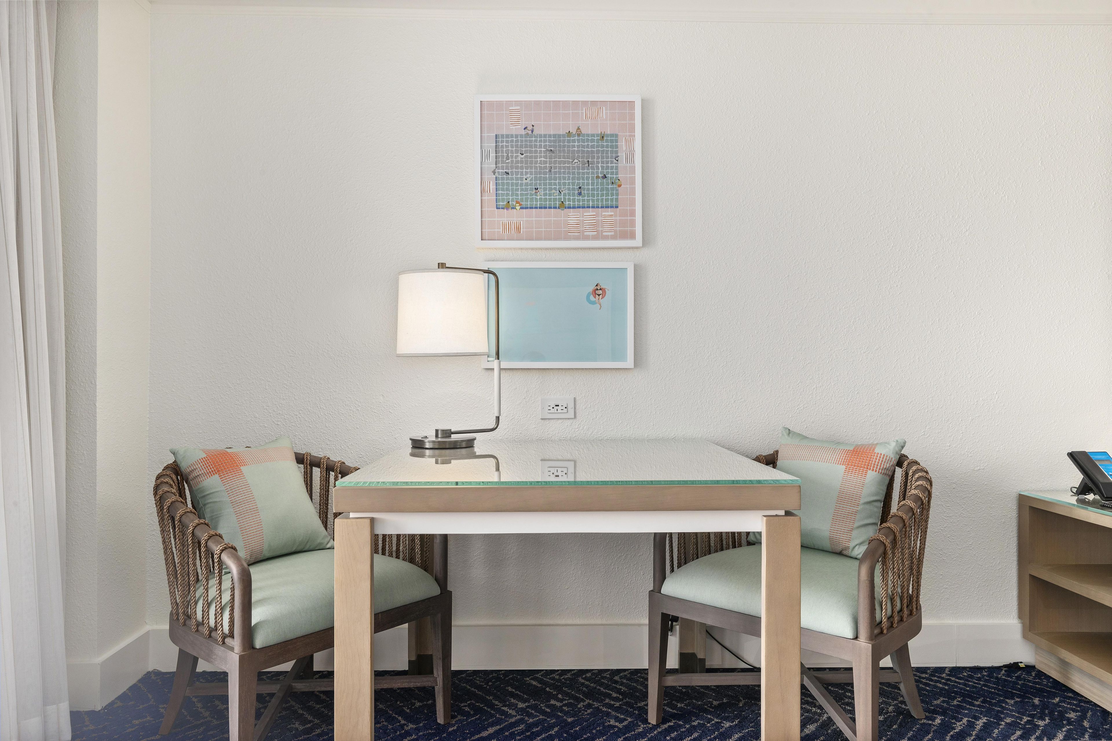 Habitación de hotel con una pequeña mesa, dos sillas, una lámpara y un cuadro en la pared.